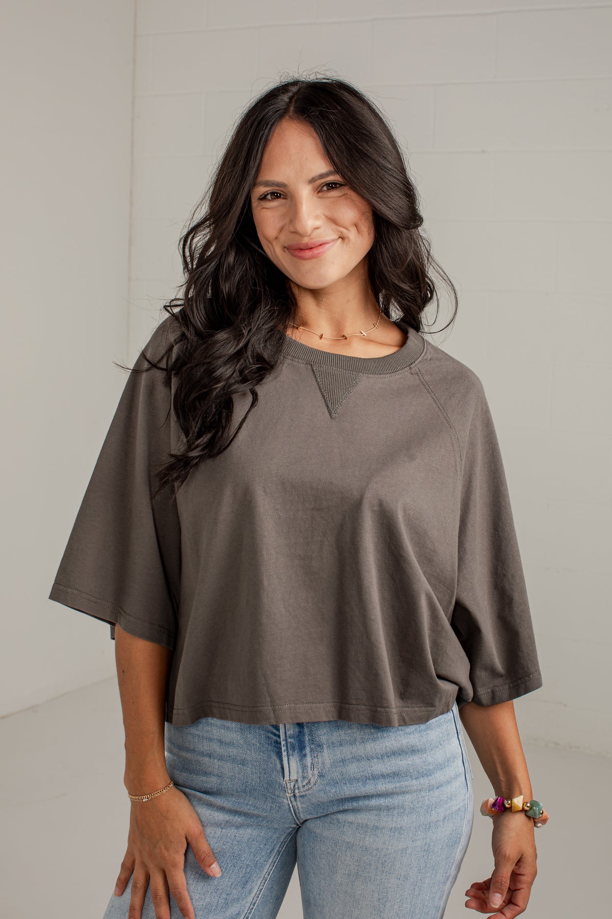 A woman with long dark hair wearing a loose-fitting, short-sleeve crewneck top in a muted taupe-brown color, paired with light-wash blue jeans, standing against a light gray wall.