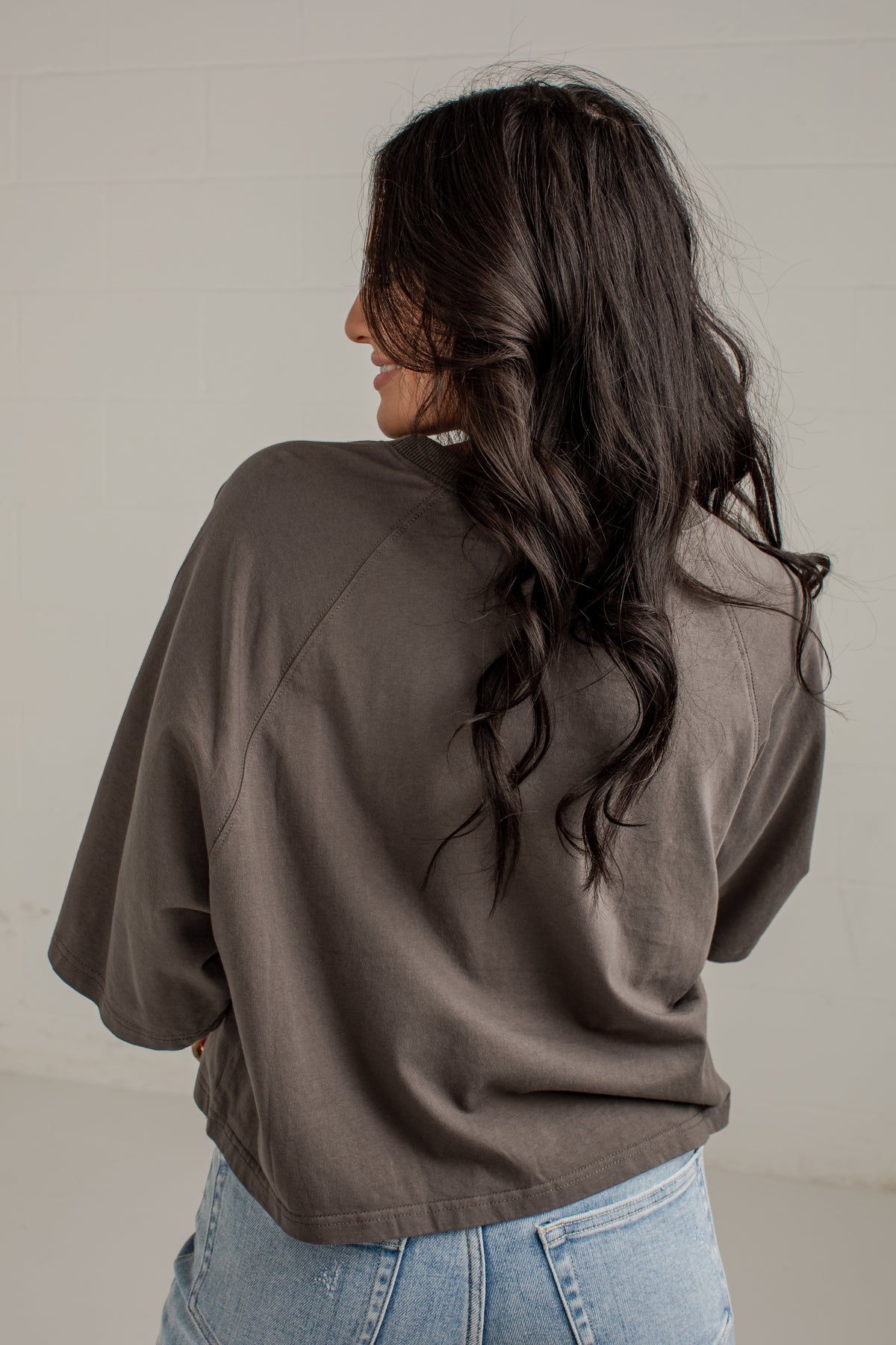 A woman with long, wavy dark hair is shown from behind, wearing an oversized dark brown short-sleeve top and light blue jeans against a light gray wall.