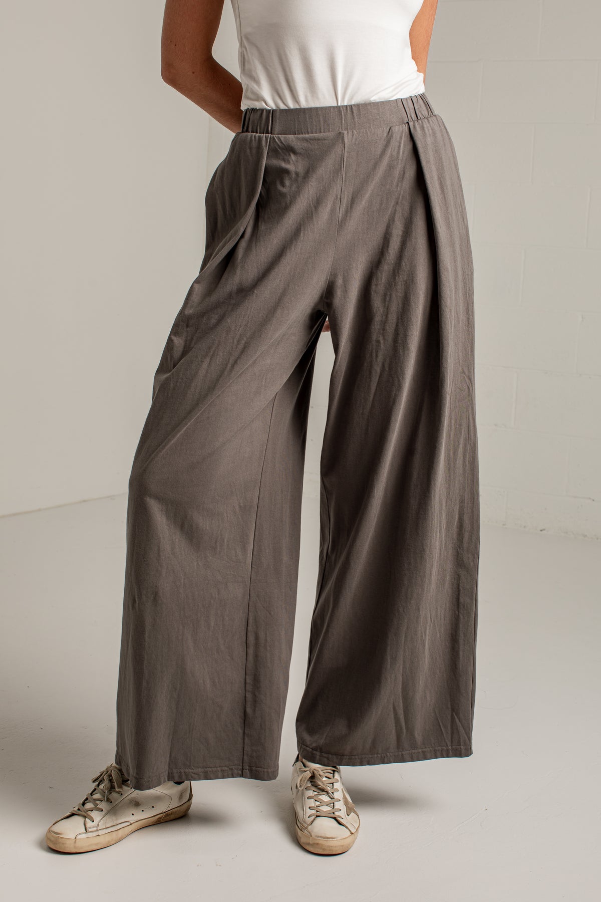 A person wearing loose, wide-leg pleated pants in a muted taupe-brown color, paired with a white sleeveless top and distressed light-colored sneakers, standing against a plain light gray wall.