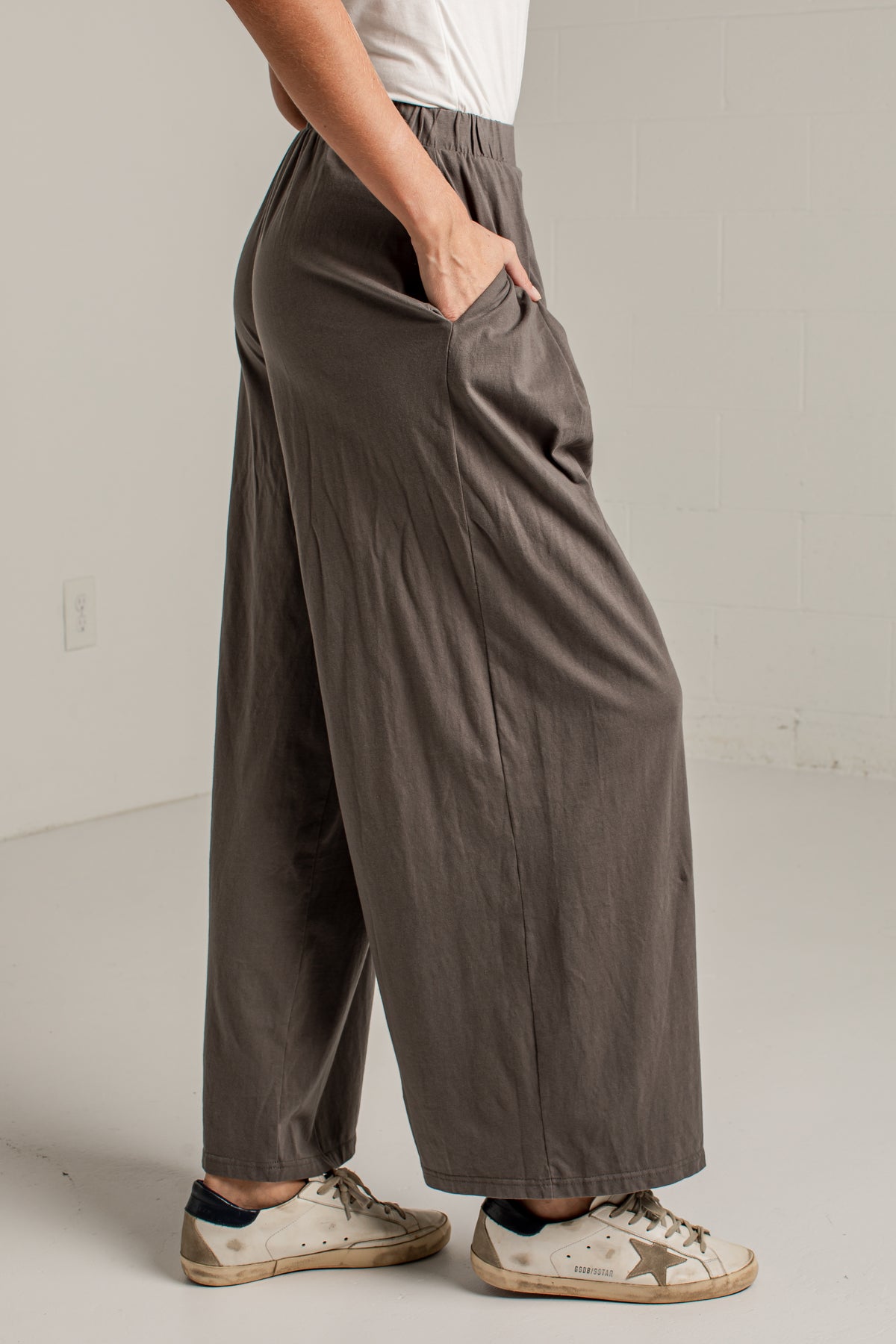 A person wearing loose, wide-leg taupe pants with side pockets, paired with a white top and distressed white sneakers, standing in a minimalist white room.