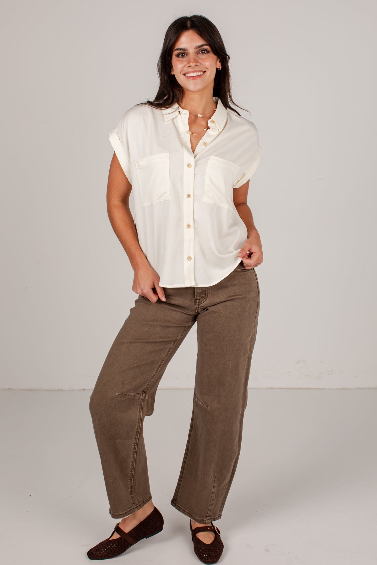 A woman modeling a short-sleeve cream button-up shirt and brown wide-leg pants against a light gray studio background.