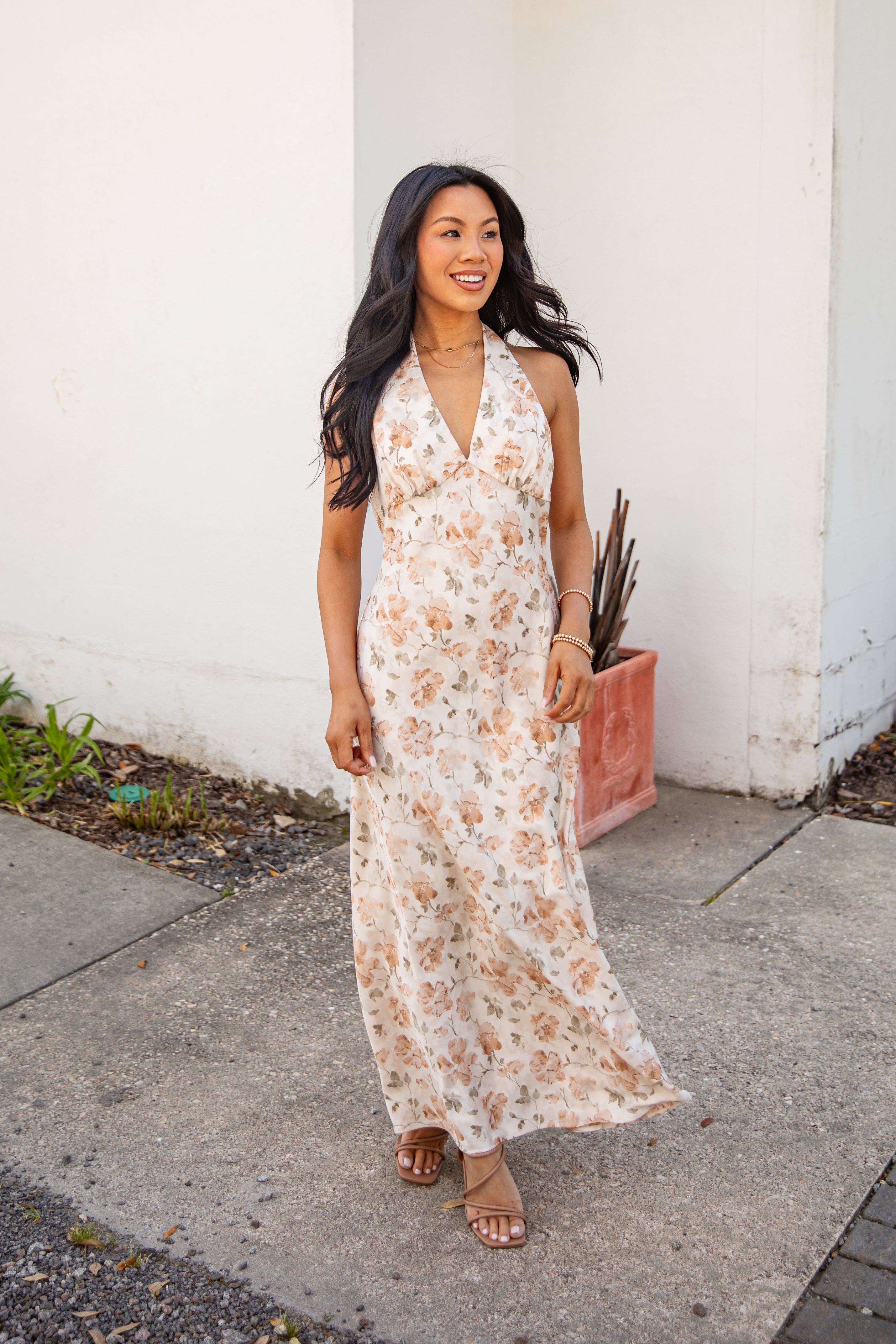 Woman in a floral dress standing on a sidewalk against a white wall.