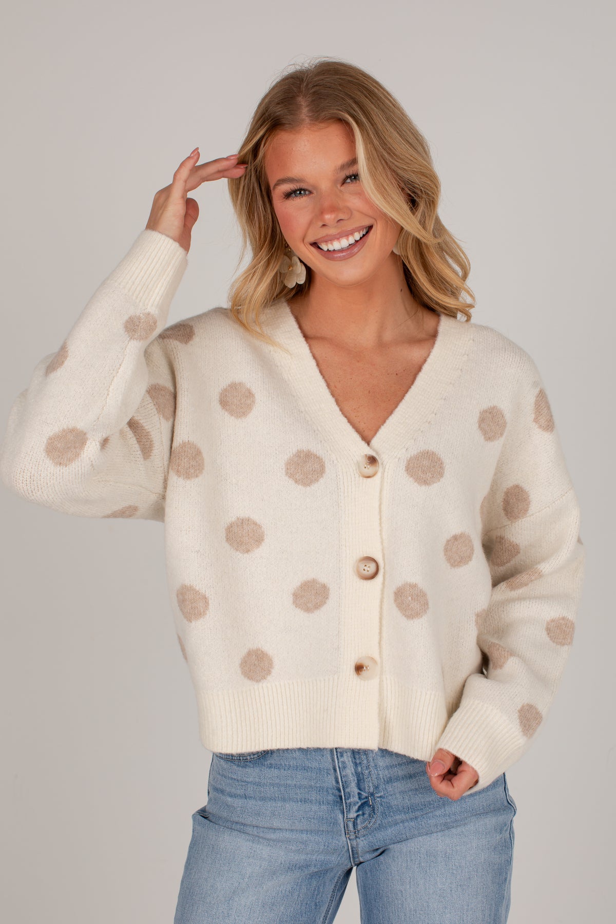 A smiling blonde woman wearing a cream knit cardigan with beige polka dots and light blue jeans, posed against a plain light gray studio background.