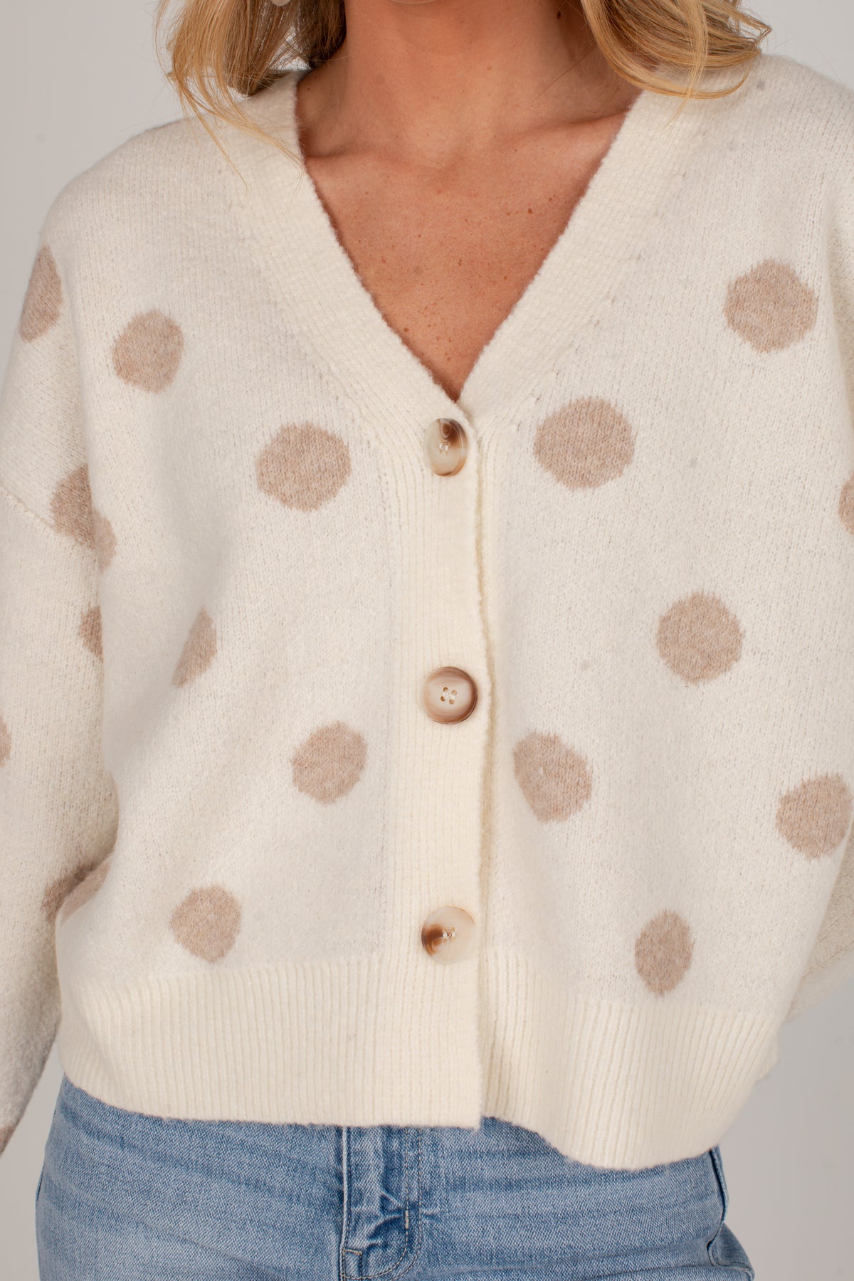 A close-up of a person wearing a cream knit cardigan with beige polka dots and light wood buttons, paired with blue jeans against a plain light gray background.