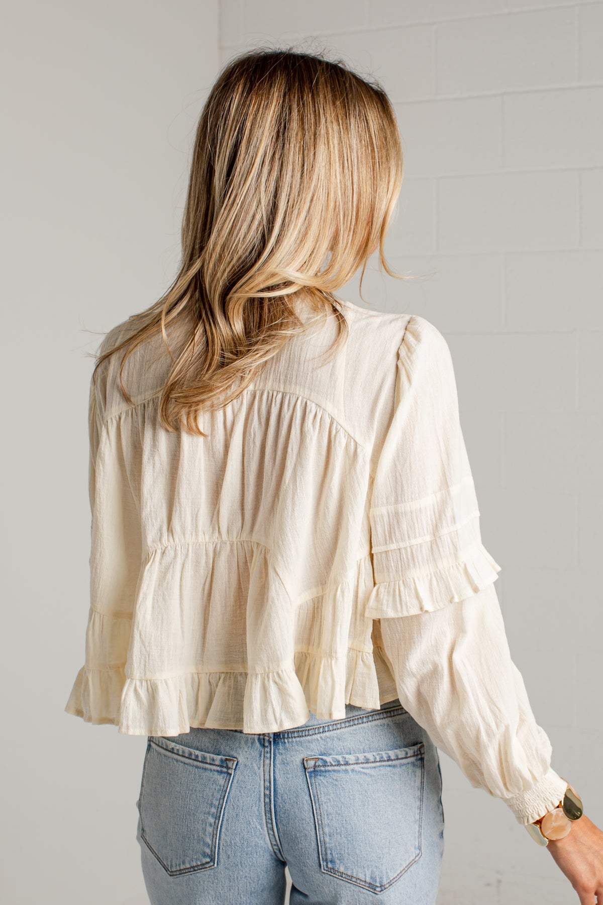 A woman viewed from behind wearing a cream-colored, long-sleeve ruffled blouse tucked into light-wash blue jeans, standing against a light gray brick wall.