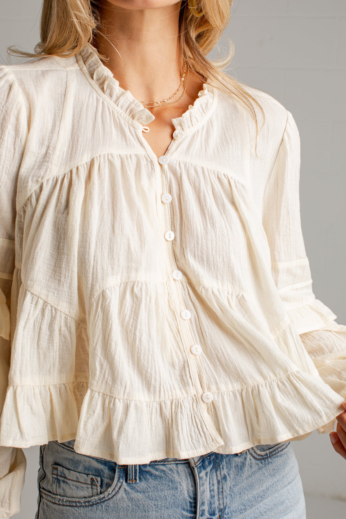 A close-up of a woman wearing a cream-colored, long-sleeve ruffled blouse with white buttons and light blue denim jeans.