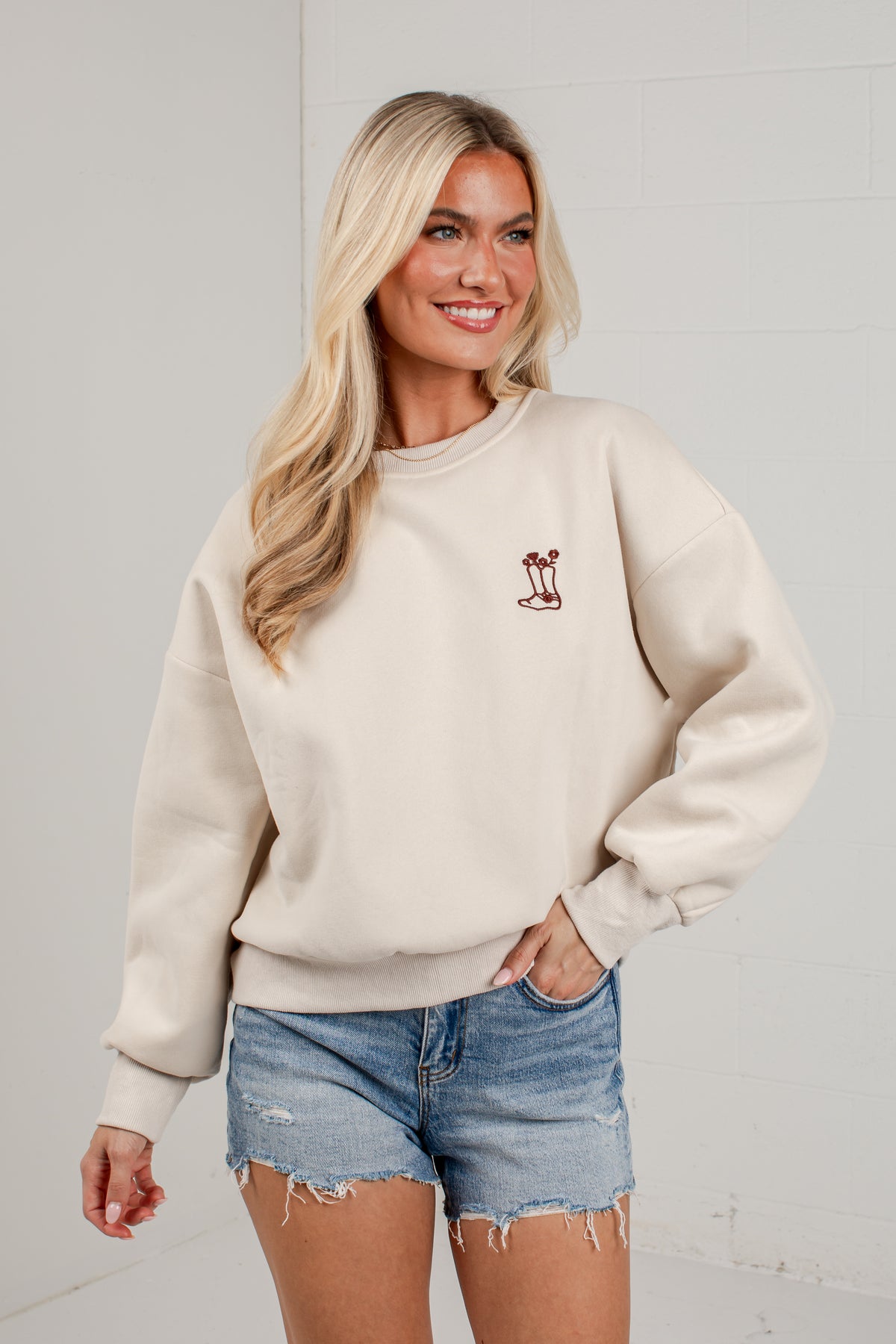 A woman modeling a cream-colored crewneck sweatshirt with a small embroidered palm tree graphic on the left chest, paired with distressed denim shorts, set against a light gray brick wall background.