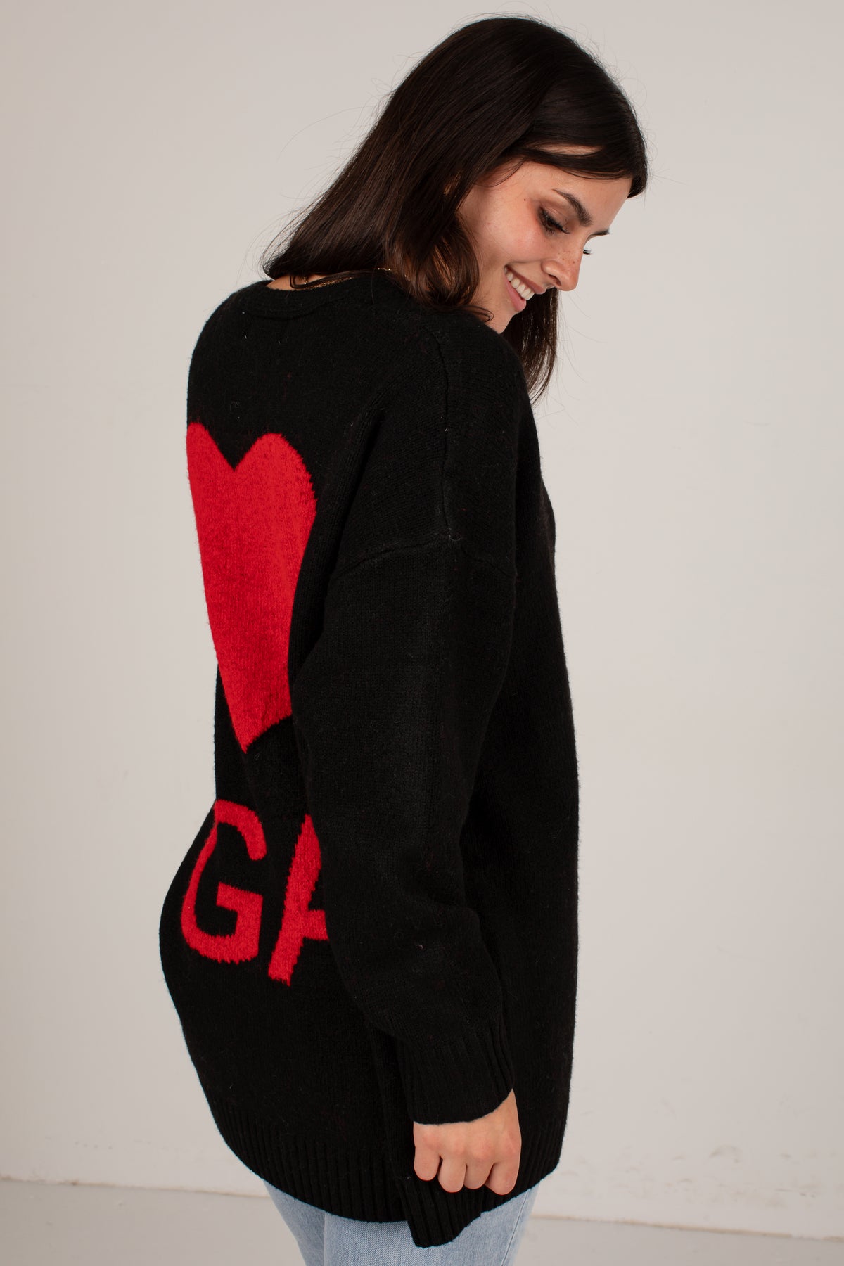 A woman wearing a black knit sweater with a large red heart and red letters on the back, photographed against a plain light wall.