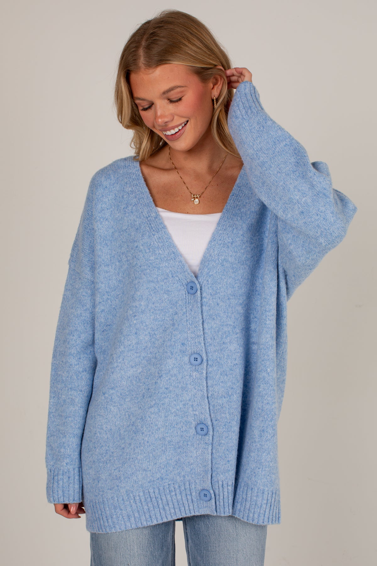 A woman wearing an oversized light blue knit cardigan with a white V-neck undershirt and light blue jeans, photographed against a plain light beige studio background.