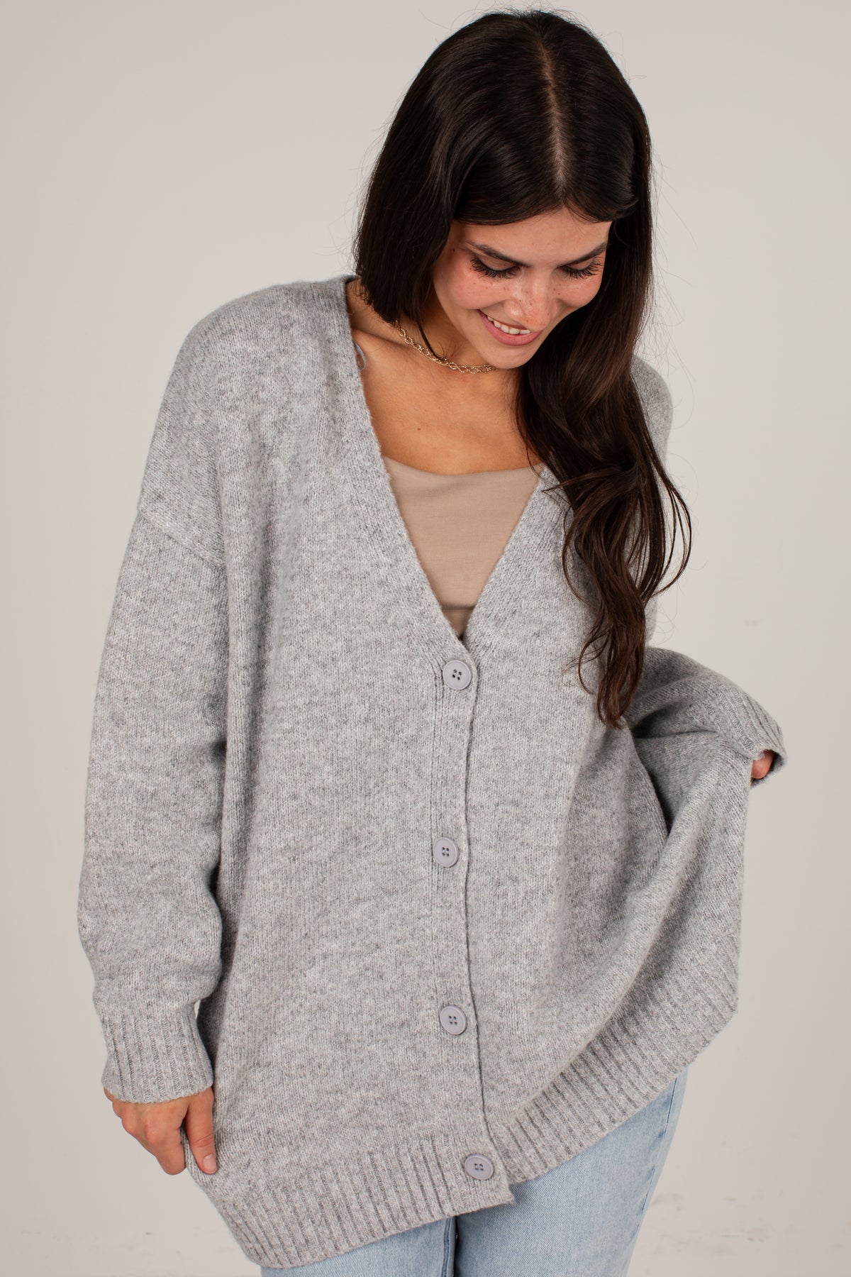 A woman wearing an oversized light gray knit cardigan with a V-neck and front buttons, layered over a beige top and light blue jeans, photographed against a plain light background.