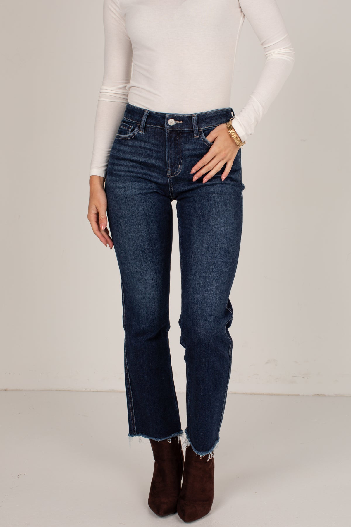 A model wearing dark blue high-waisted straight-leg jeans with a raw frayed hem, styled with a white long-sleeve top and brown suede ankle boots against a plain off-white studio background.