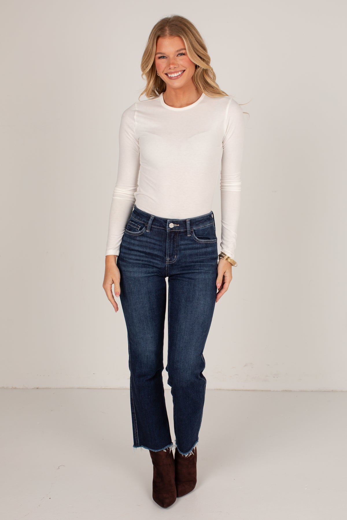 A woman modeling a white long-sleeve crewneck top and dark blue straight-leg jeans with a raw hem, paired with brown ankle boots, against a plain off-white studio background.