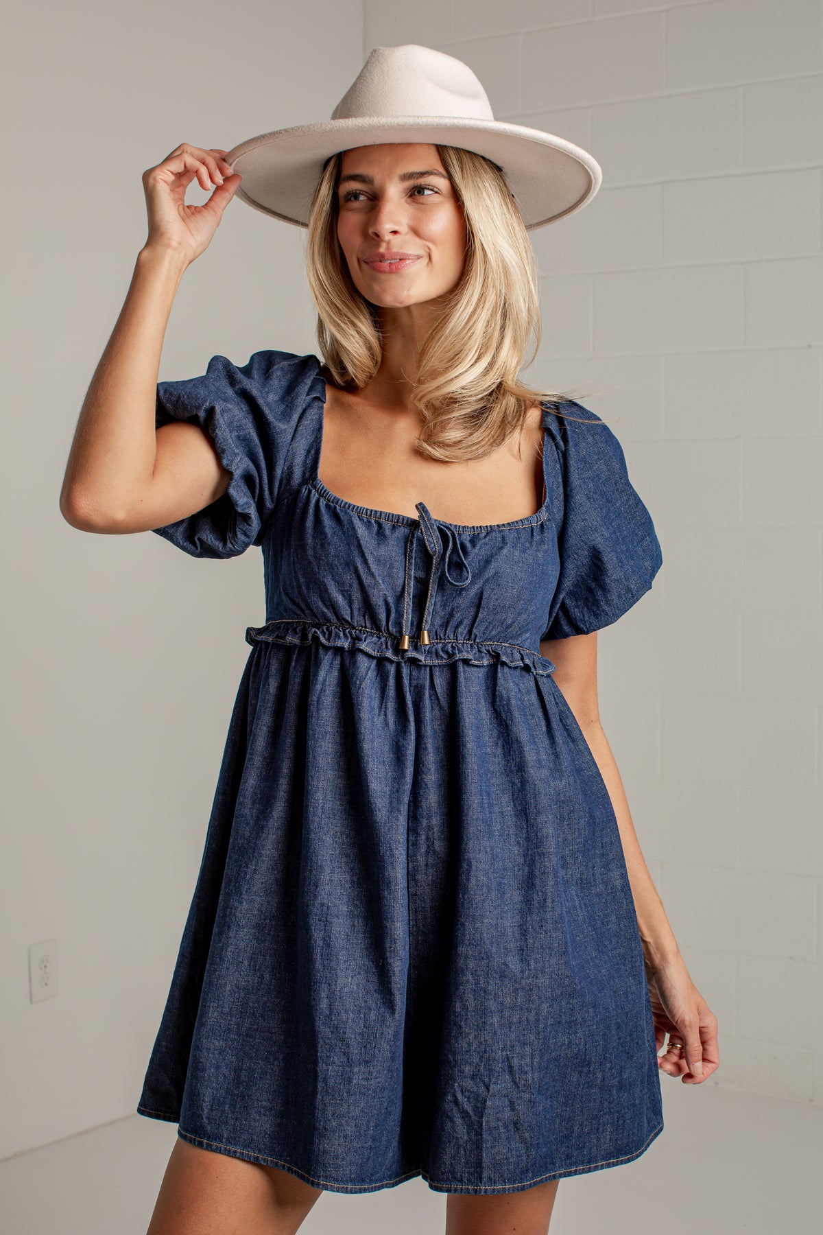 A woman wearing a blue denim dress with puff sleeves, a square neckline, and a drawstring waist, paired with a wide-brimmed white hat, posing against a light gray wall.
