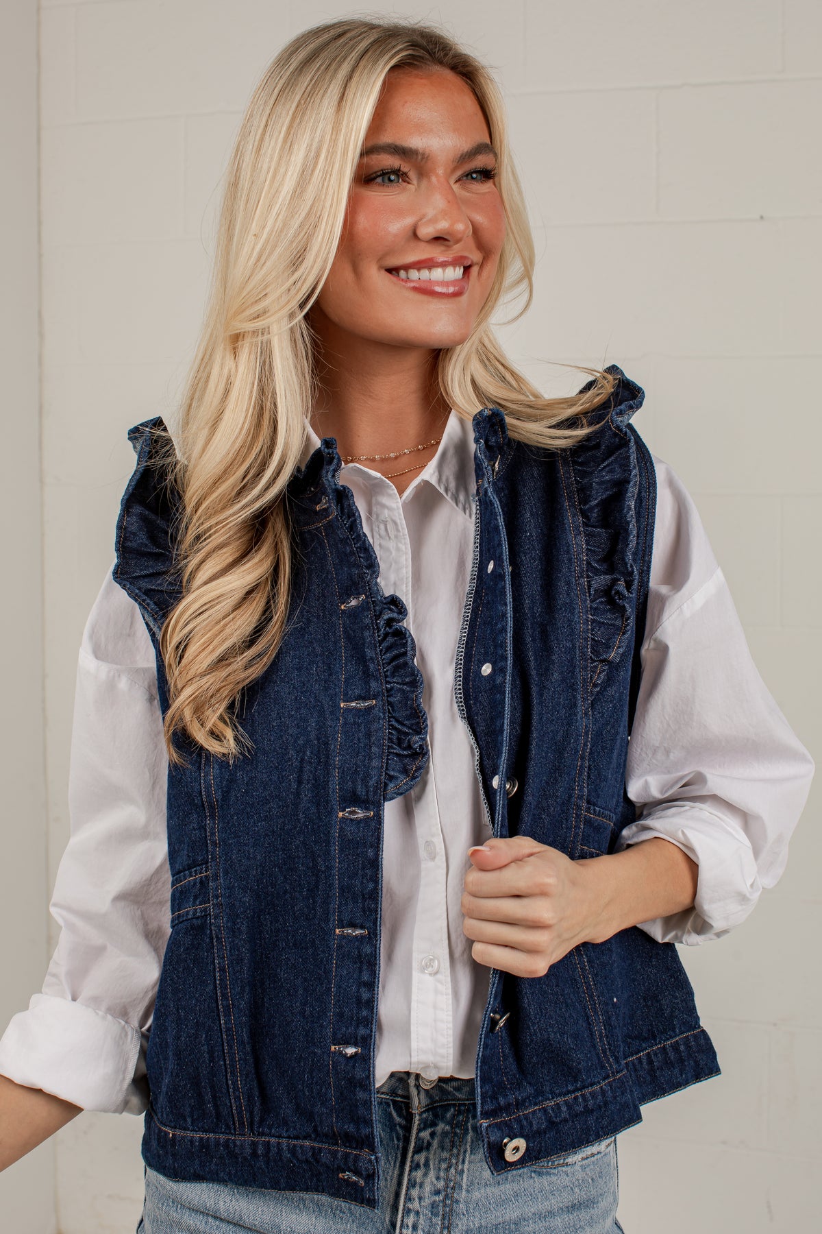 A smiling blonde woman wearing a dark blue denim vest with ruffle trim over a white button-down shirt and light-wash jeans, standing against a light neutral wall.