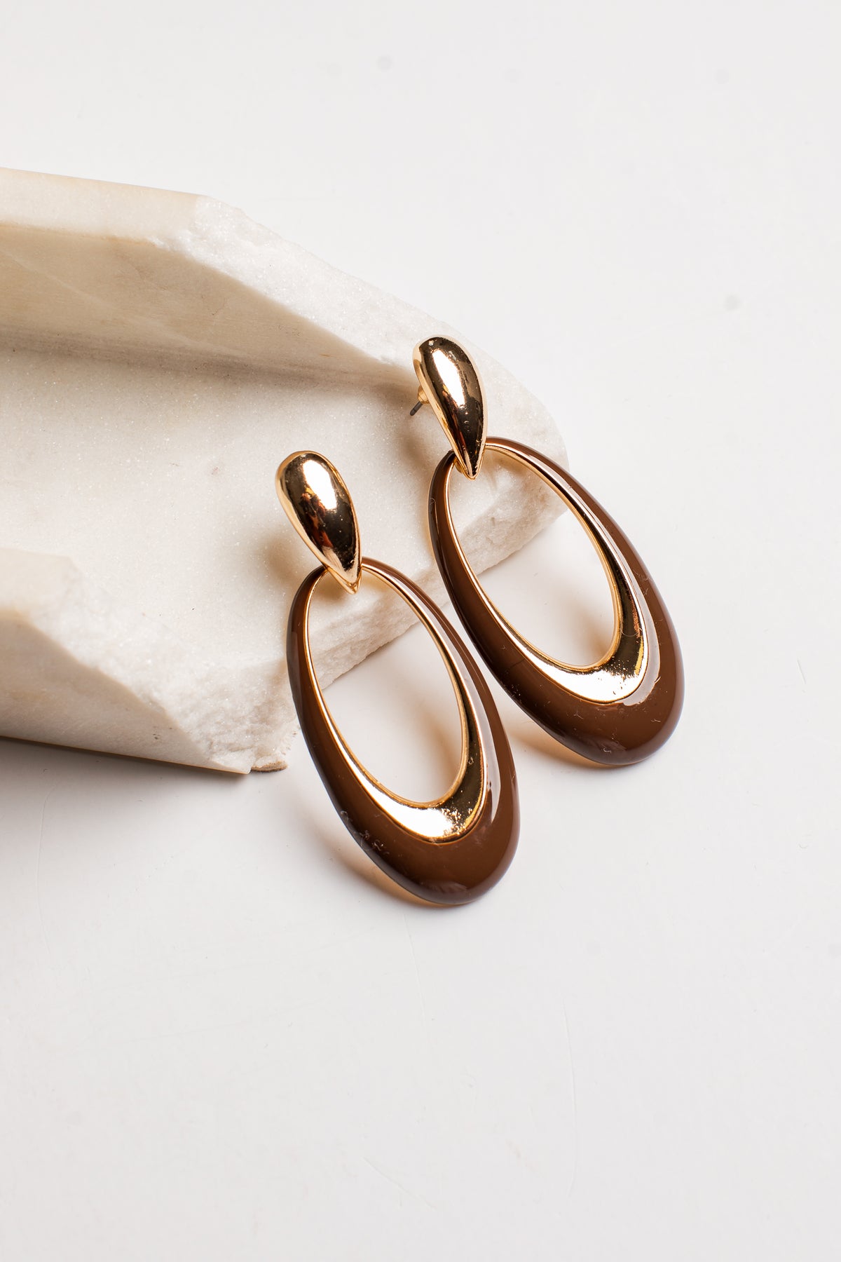 A pair of gold and brown oval drop earrings displayed on a light beige stone prop against a clean white background.