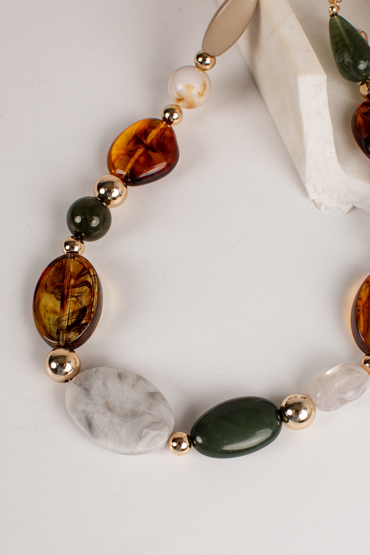A close-up of a beaded necklace featuring large marbled amber-brown beads, a white pearl, a dark green stone, a white marbled stone, and polished gold spacer beads, arranged on a light neutral background.