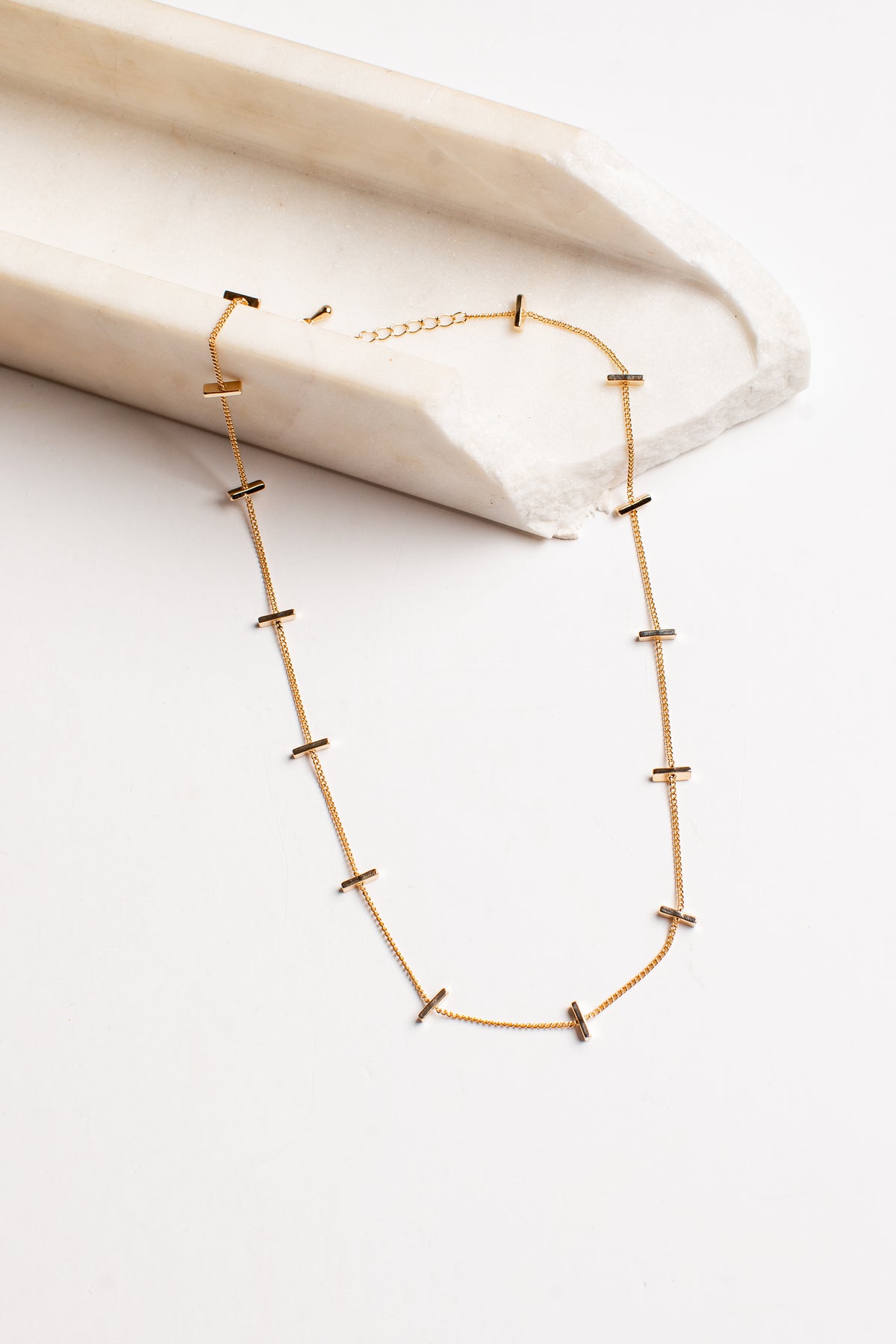 A delicate gold chain necklace with small, spaced cross-shaped links, displayed on a white surface next to a cream-colored stone block.
