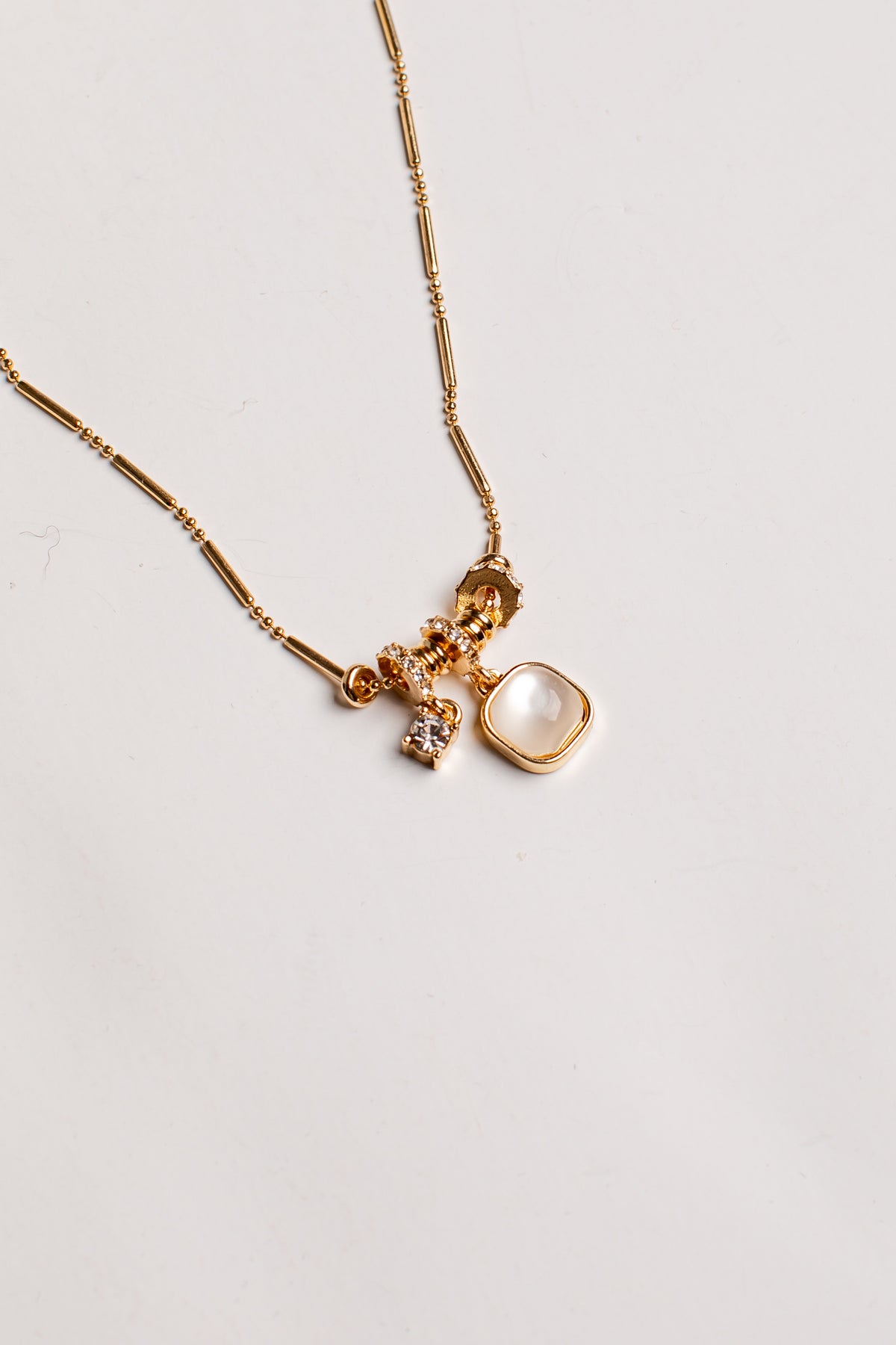 A gold chain necklace featuring a multi-charm pendant with a large square cream-colored stone, a small faceted gold bead, and a tiny clear crystal charm, displayed on a clean off-white background.