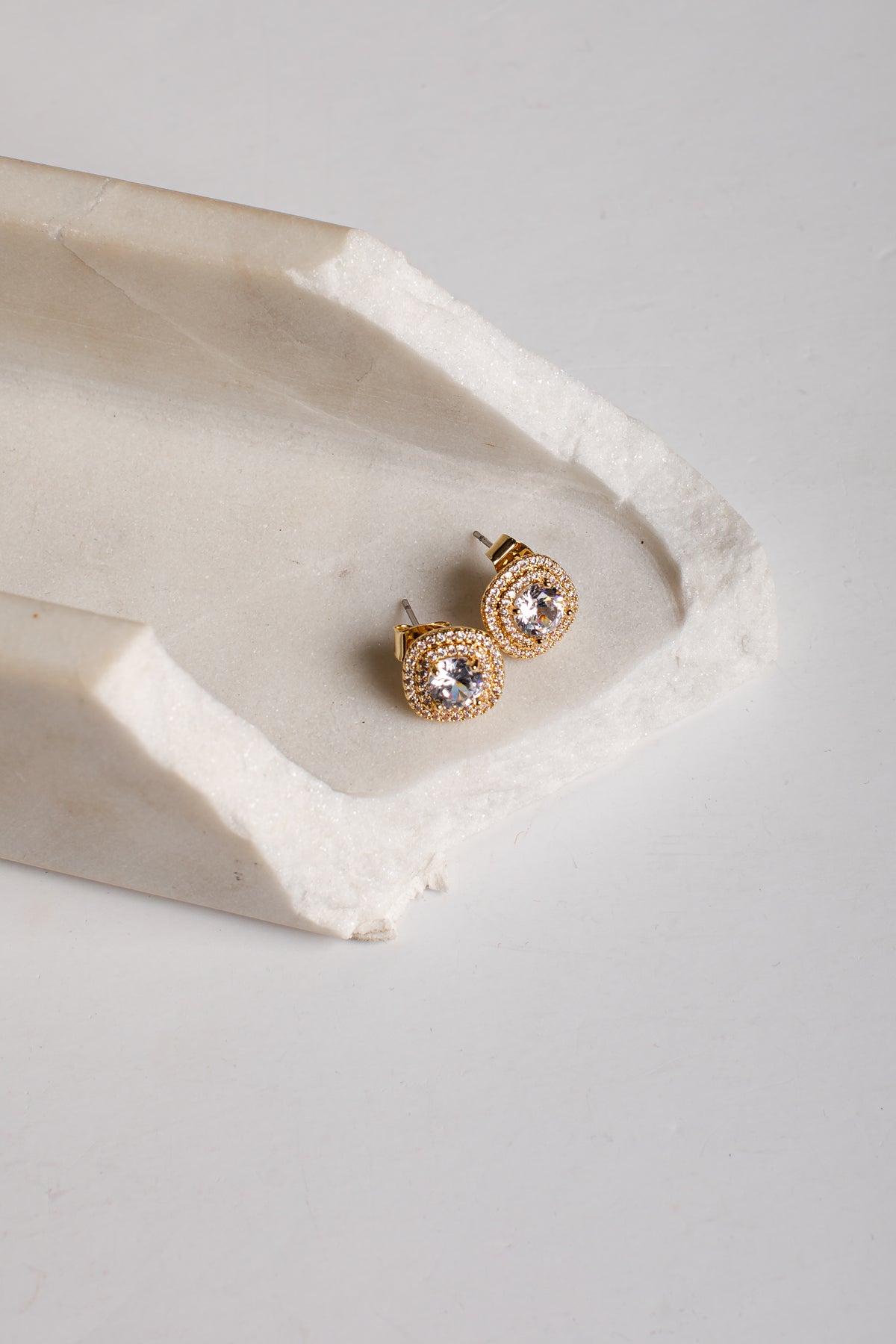A pair of gold stud earrings with clear round gemstones, displayed on a textured white stone tray against a light gray background.