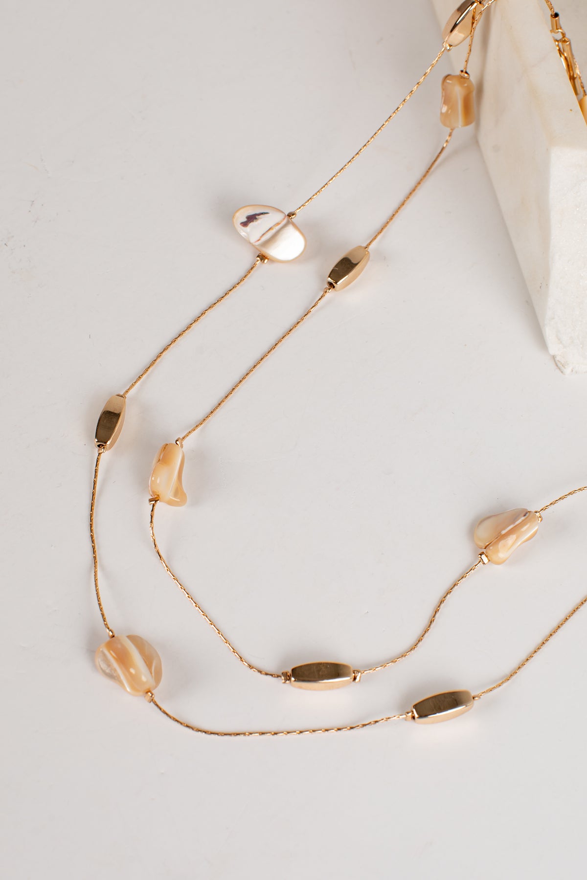 A gold-toned multi-strand necklace featuring irregular beige shell beads and polished oval bar accents, styled on a light gray surface with a white marble prop.