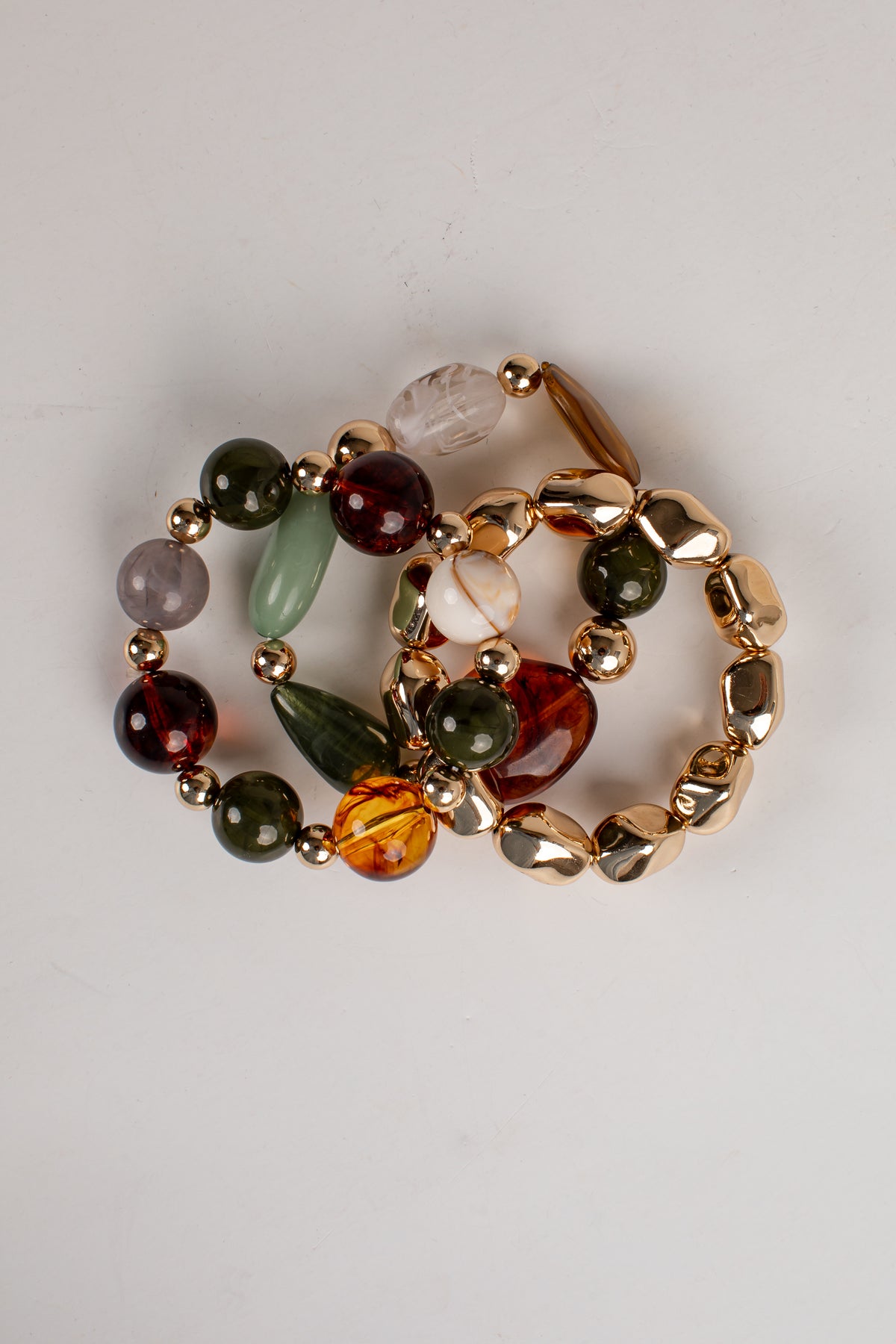 Three beaded stretch bracelets with gold-tone metal links and assorted gemstone beads in green, amber, brown, purple, and white, arranged on a light gray background.
