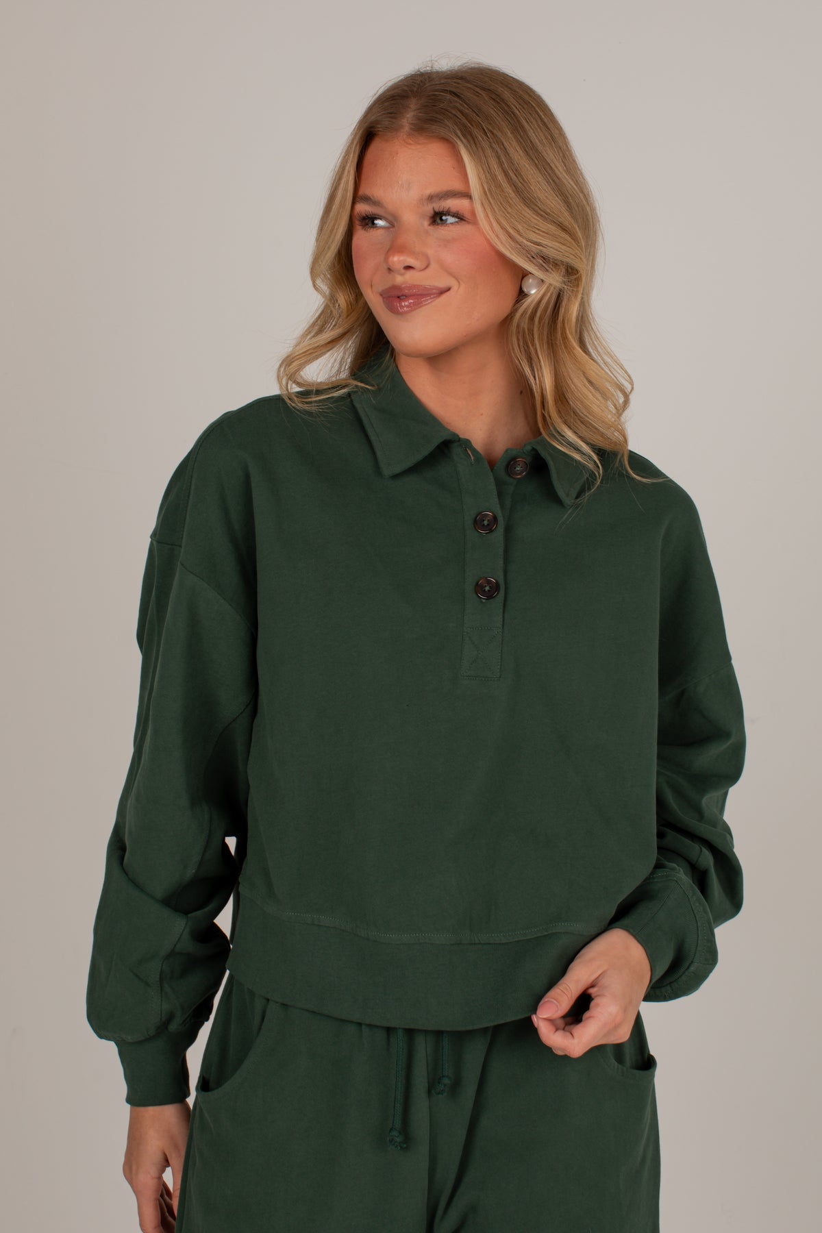 A woman wearing a dark forest green long-sleeve collared pullover top with a three-button placket, paired with matching green drawstring pants, against a plain light gray studio background.