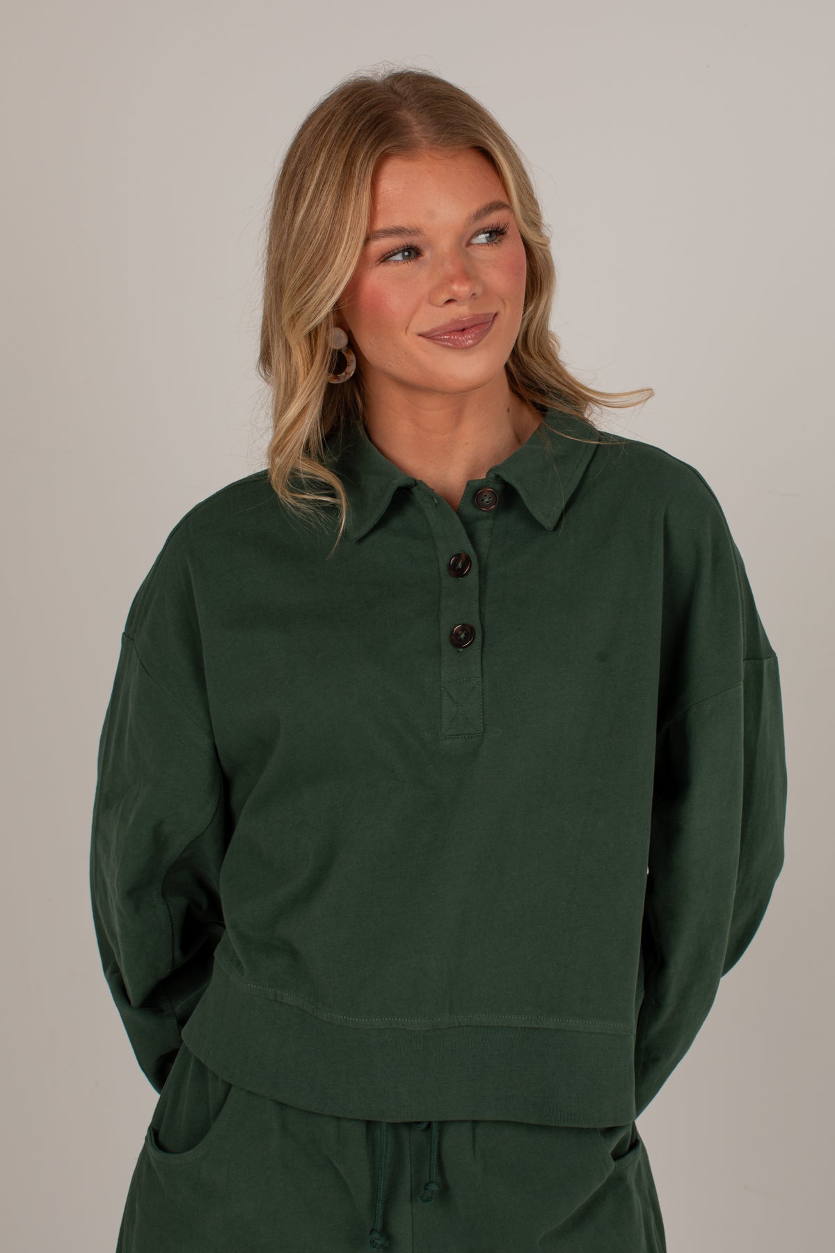 A woman wearing a dark forest green long-sleeve polo sweatshirt with a buttoned collar, paired with matching green sweatpants, set against a plain light gray studio background.