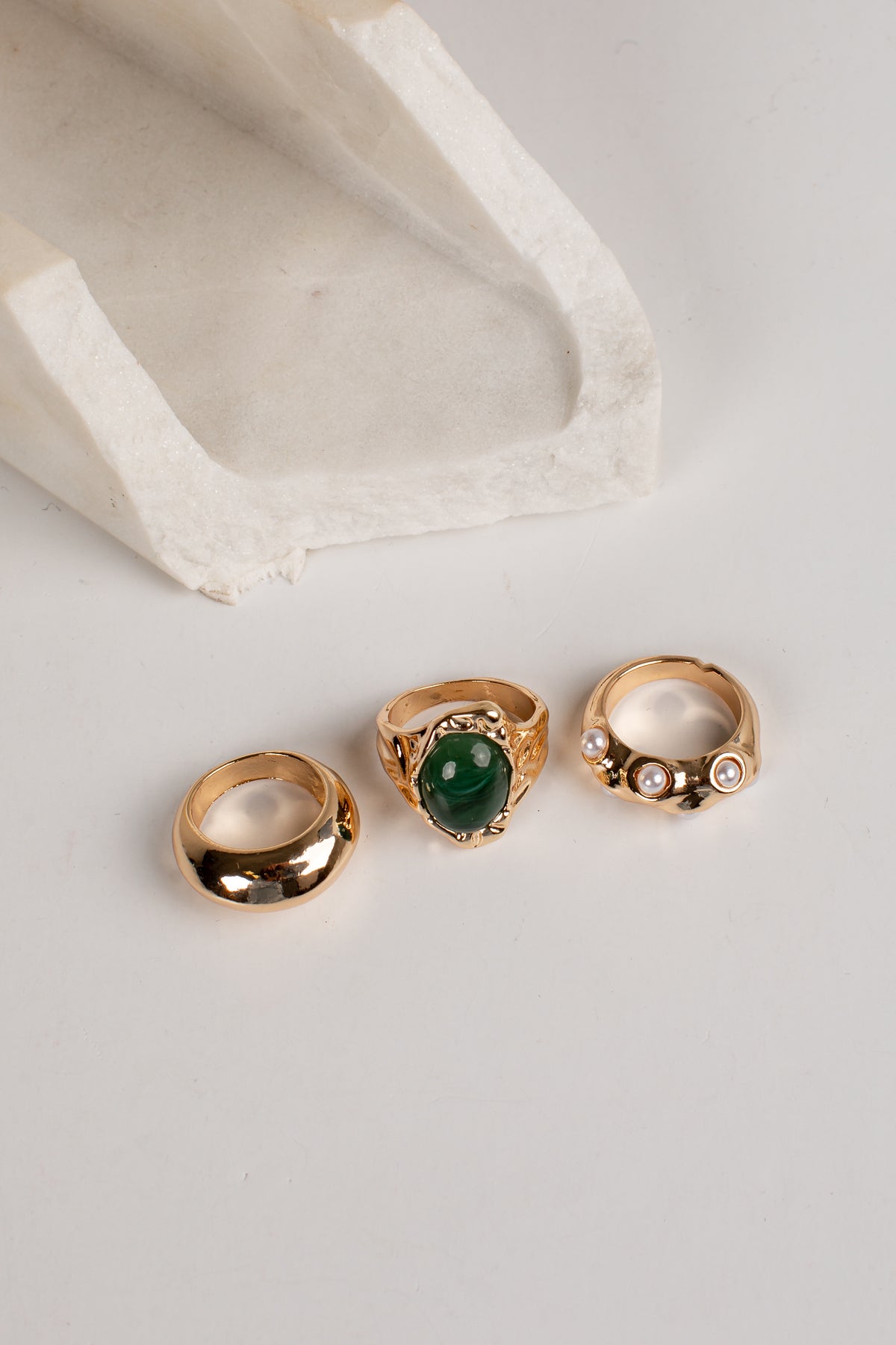 Three gold rings displayed on a light gray surface with a white textured stone prop. The left ring is a smooth polished band, the center ring features a large oval green stone, and the right ring has a textured band with small clear stones.
