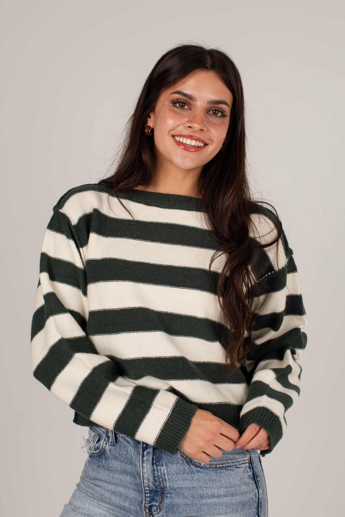 A smiling woman with long dark hair wearing a dark green and cream horizontal striped knit sweater and light blue jeans, standing against a plain light gray studio background.
