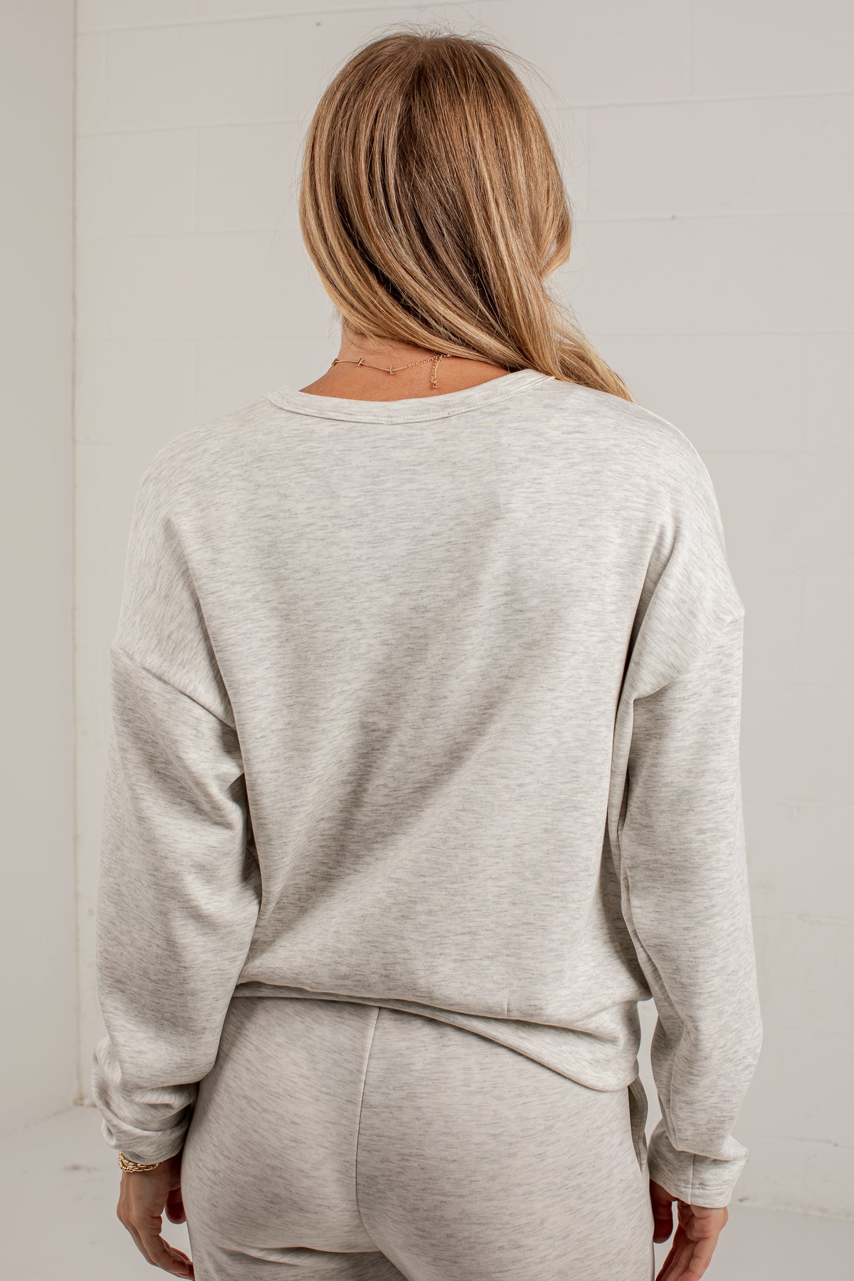 A woman viewed from behind wearing a light heather gray crewneck sweatshirt and matching sweatpants against a white paneled wall.
