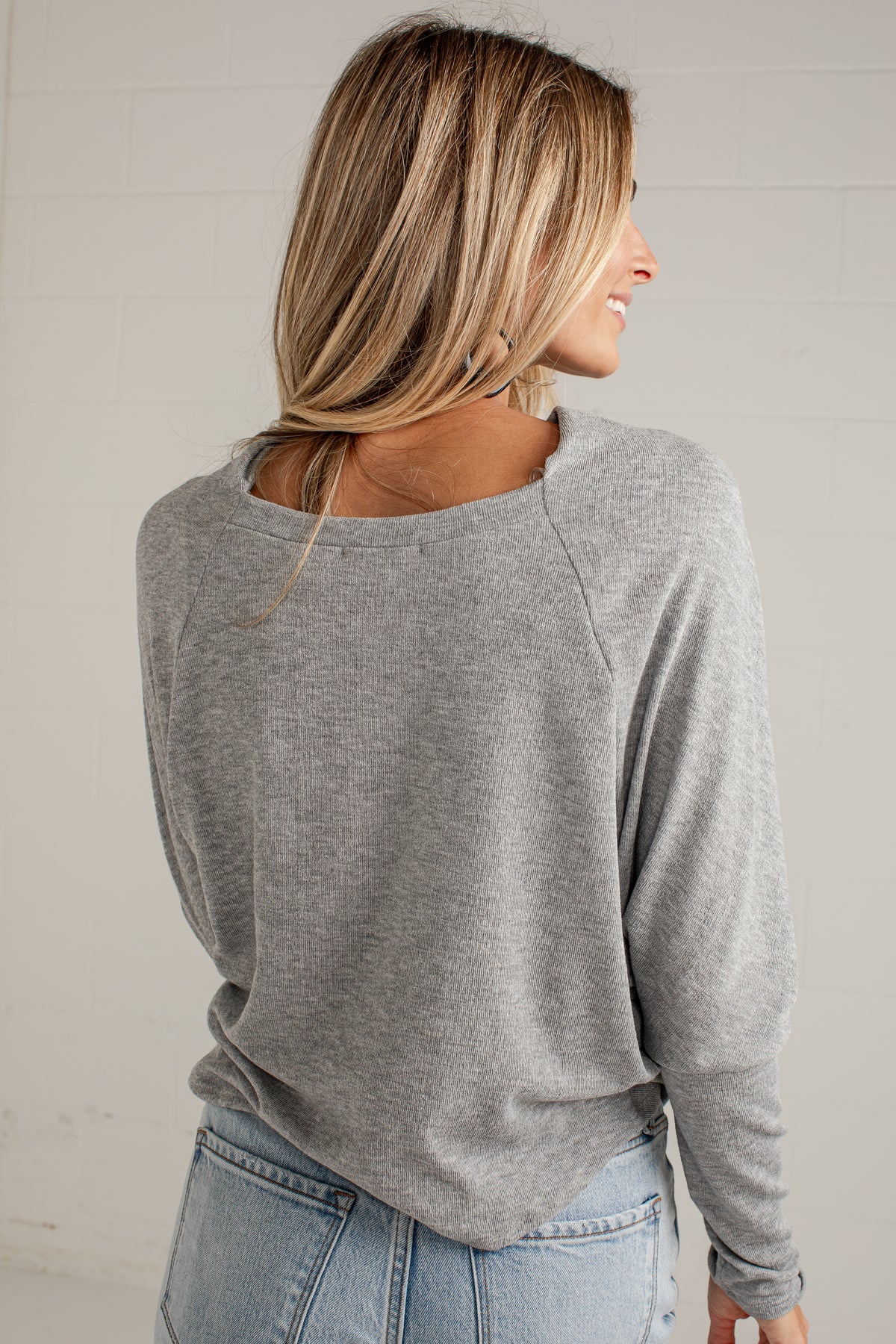 A woman shown from the back wearing a heather gray long-sleeve raglan top and light blue jeans, standing against a light off-white brick wall.