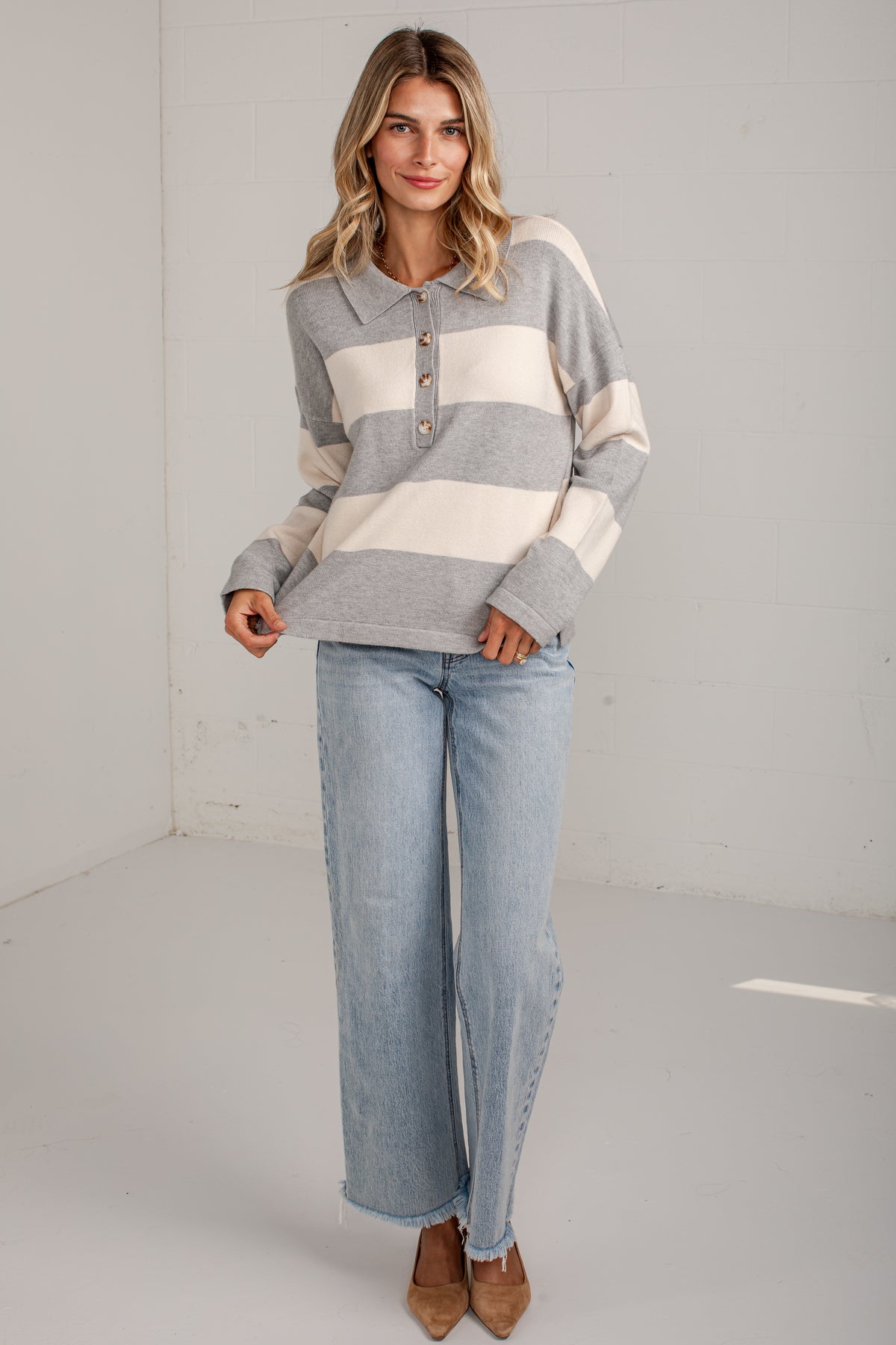 A woman modeling a gray and cream striped long-sleeve henley sweater with light-wash wide-leg jeans and brown pointed-toe heels, standing in a bright white studio corner.