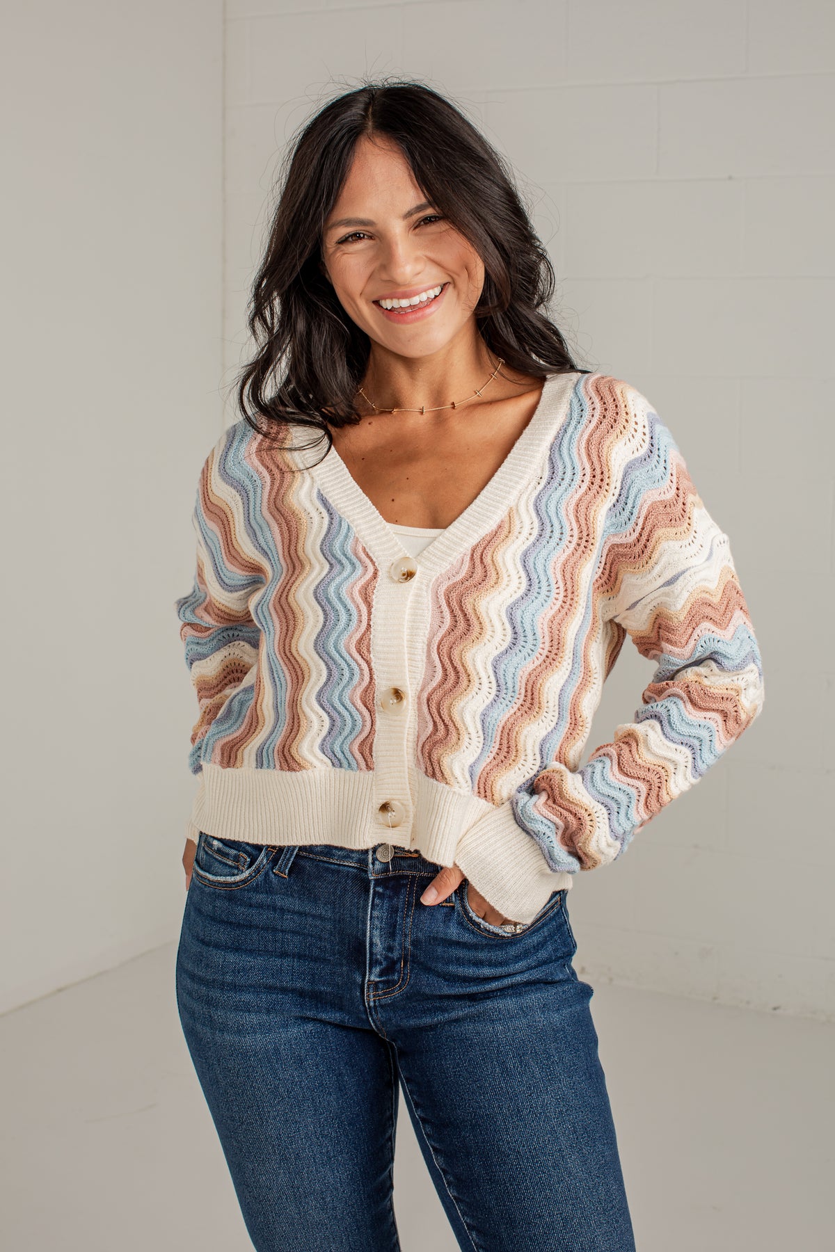 A woman wearing a cream-colored knit cardigan with wavy stripes in light blue, dusty pink, and tan, paired with dark blue jeans, standing against a light gray wall.