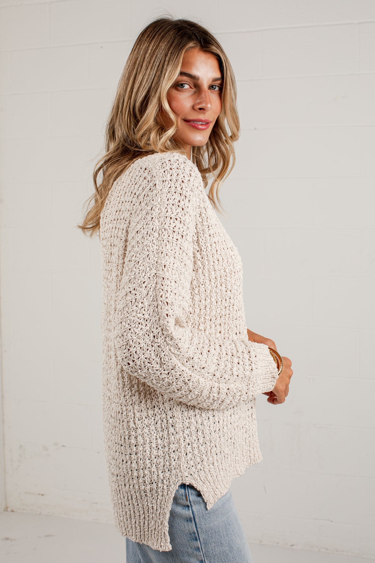 A woman wearing a chunky, cream-colored open-knit sweater and light blue jeans, standing against a white paneled wall.