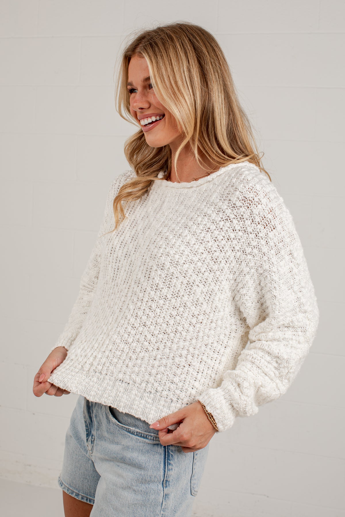 A woman wearing a chunky knit cream-colored sweater and light blue denim shorts, smiling and posing against a light gray wall.