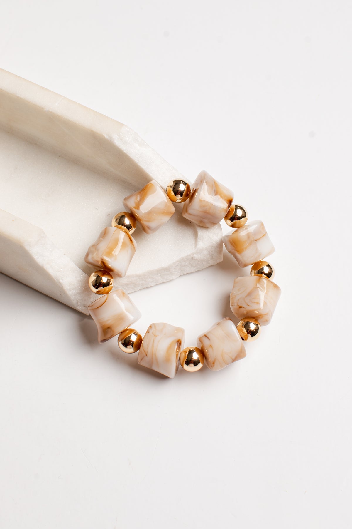 A beaded stretch bracelet featuring cream and light brown marbled square beads separated by small polished gold metallic beads, displayed on a white paper towel roll against a white background.