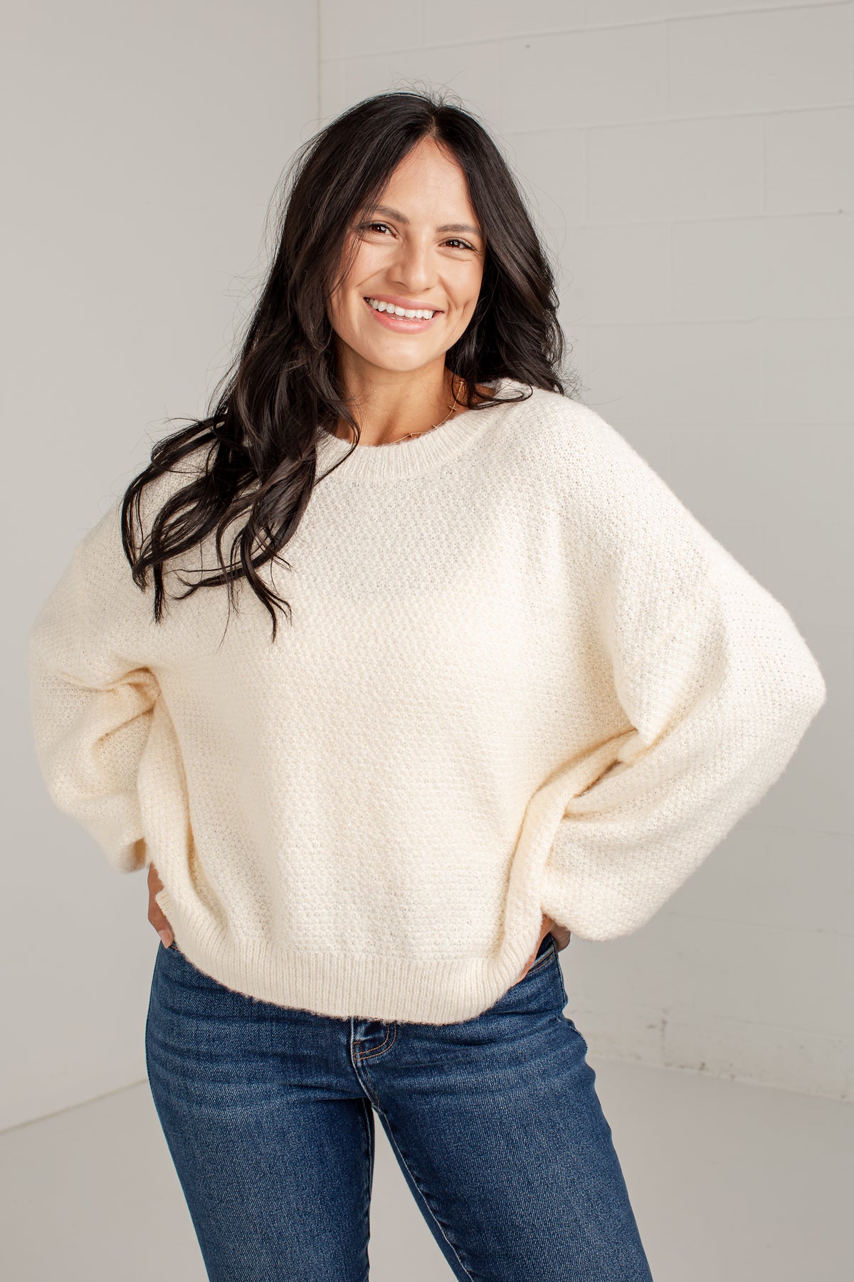 A smiling woman with long dark hair wears a cream-colored, textured bubble-sleeve knit sweater and blue jeans, standing against a light gray wall.