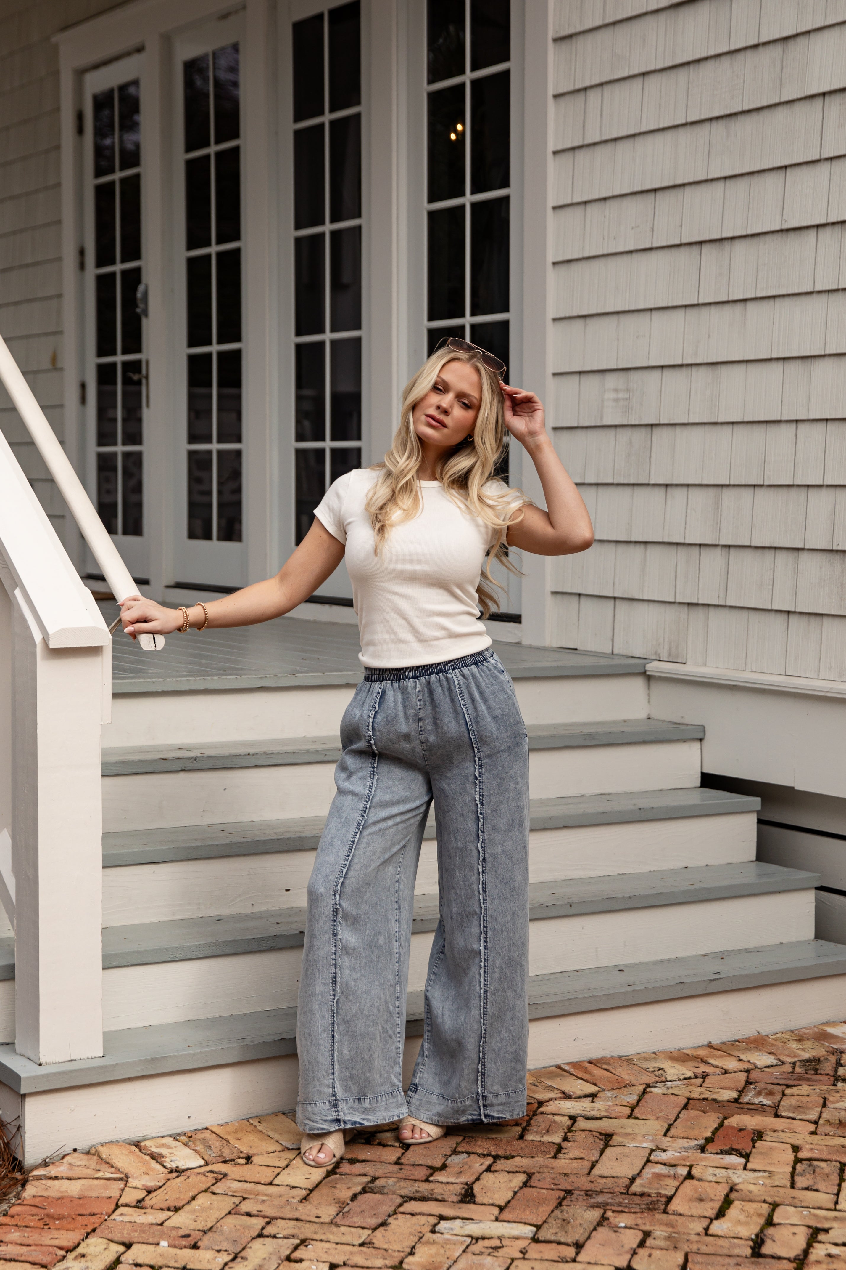 Woman standing on a set of stairs wearing a white top and blue jeans, with a neutral-colored house in the background.