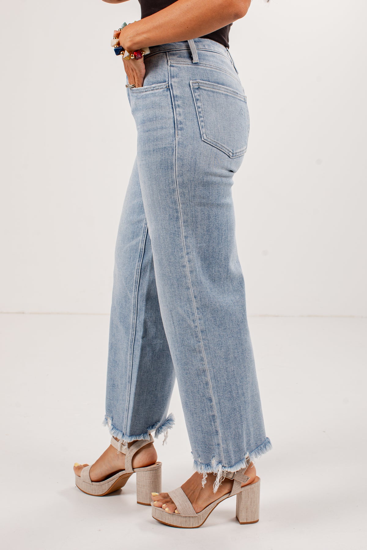 A person wearing light-wash blue wide-leg jeans with a raw frayed hem, paired with beige platform block-heel sandals, shown from the waist down against a plain white studio background.