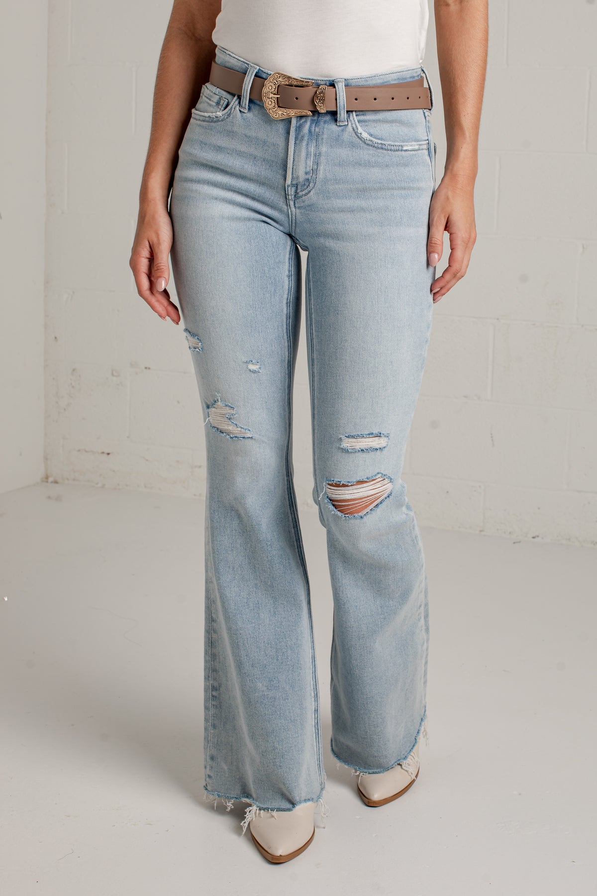 A person wearing light-wash blue distressed flare jeans with a brown belt, standing against a white studio wall.