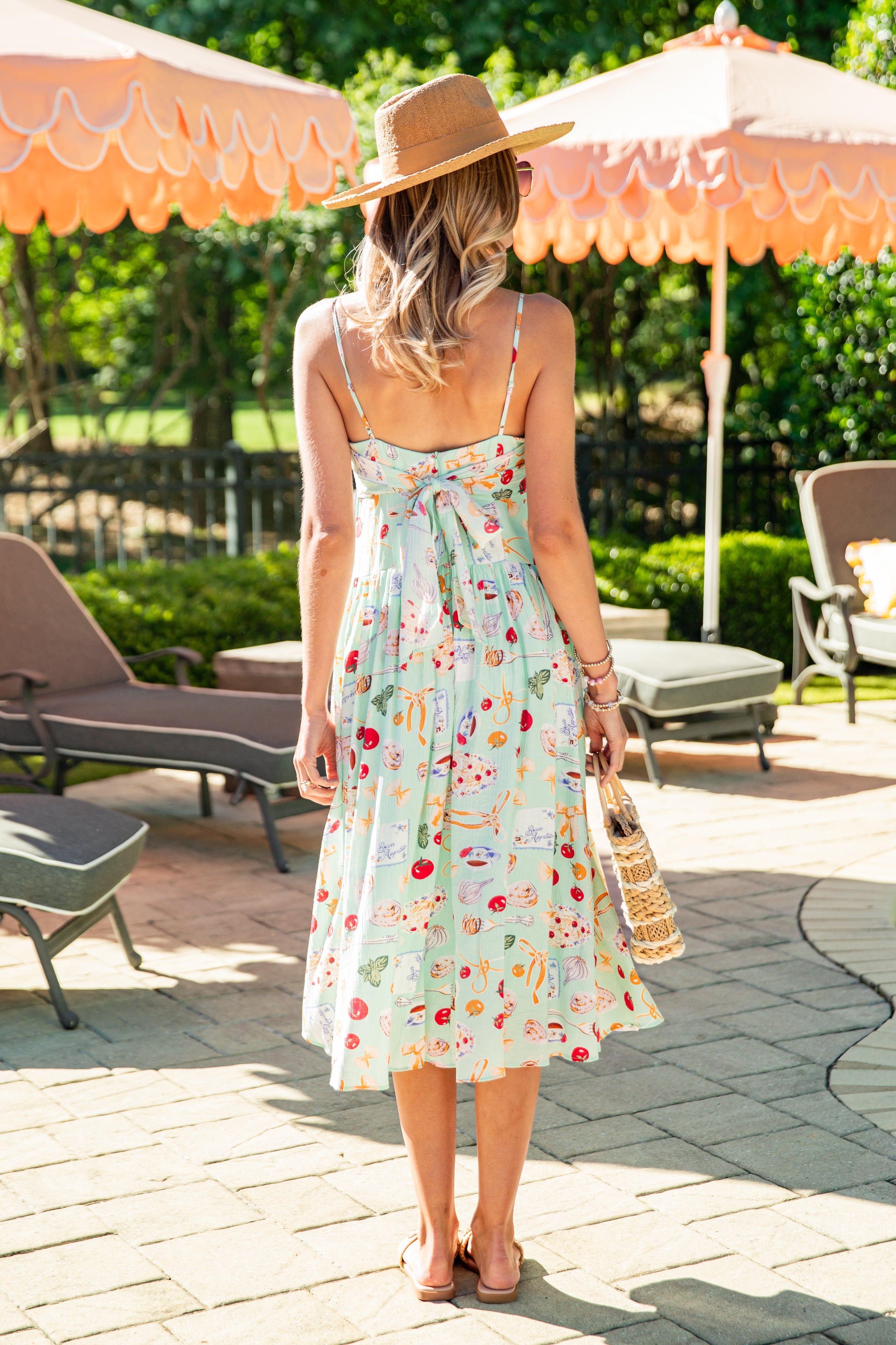 Woman in a floral dress and hat standing in a sunlit outdoor setting with umbrellas and chairs.