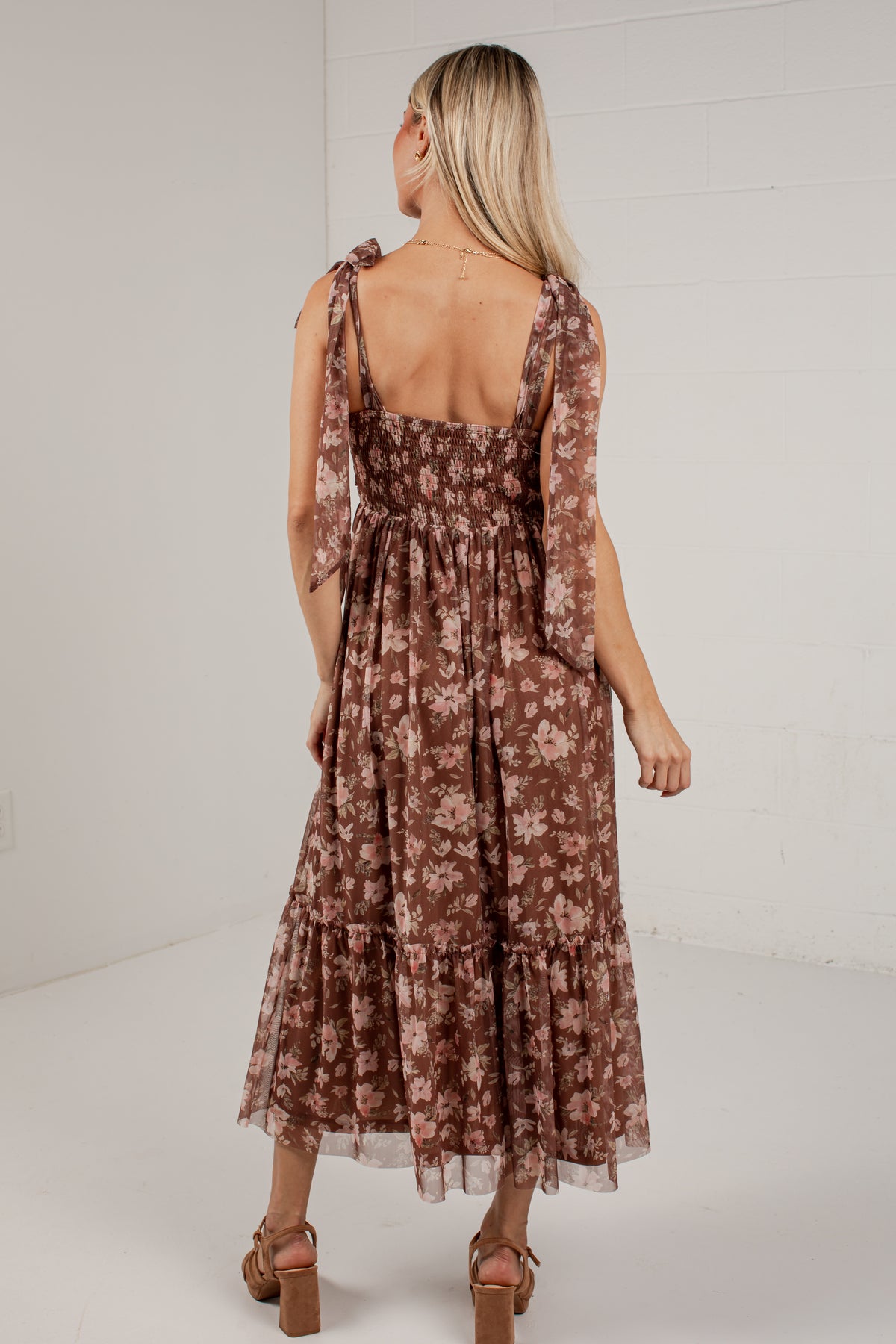 A woman wearing a brown tiered midi dress with a floral pattern, shown from the back against a white studio background.