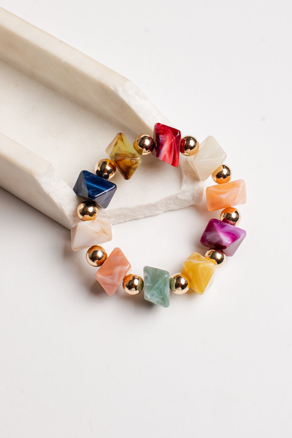 A multicolor beaded stretch bracelet with faceted gemstone-like beads in various colors and small gold spacer beads, displayed on a white surface next to a light wood block.