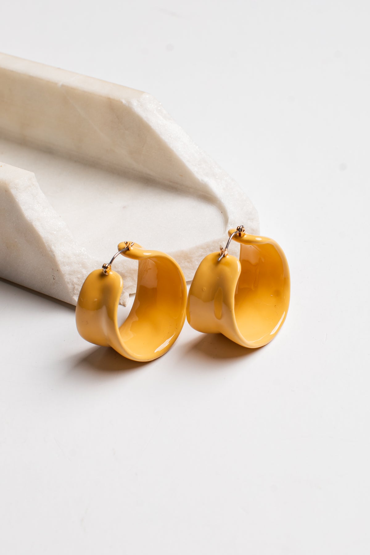 A pair of chunky, glossy mustard yellow hoop earrings with small gold posts, displayed on a white marble prop against a clean white background.