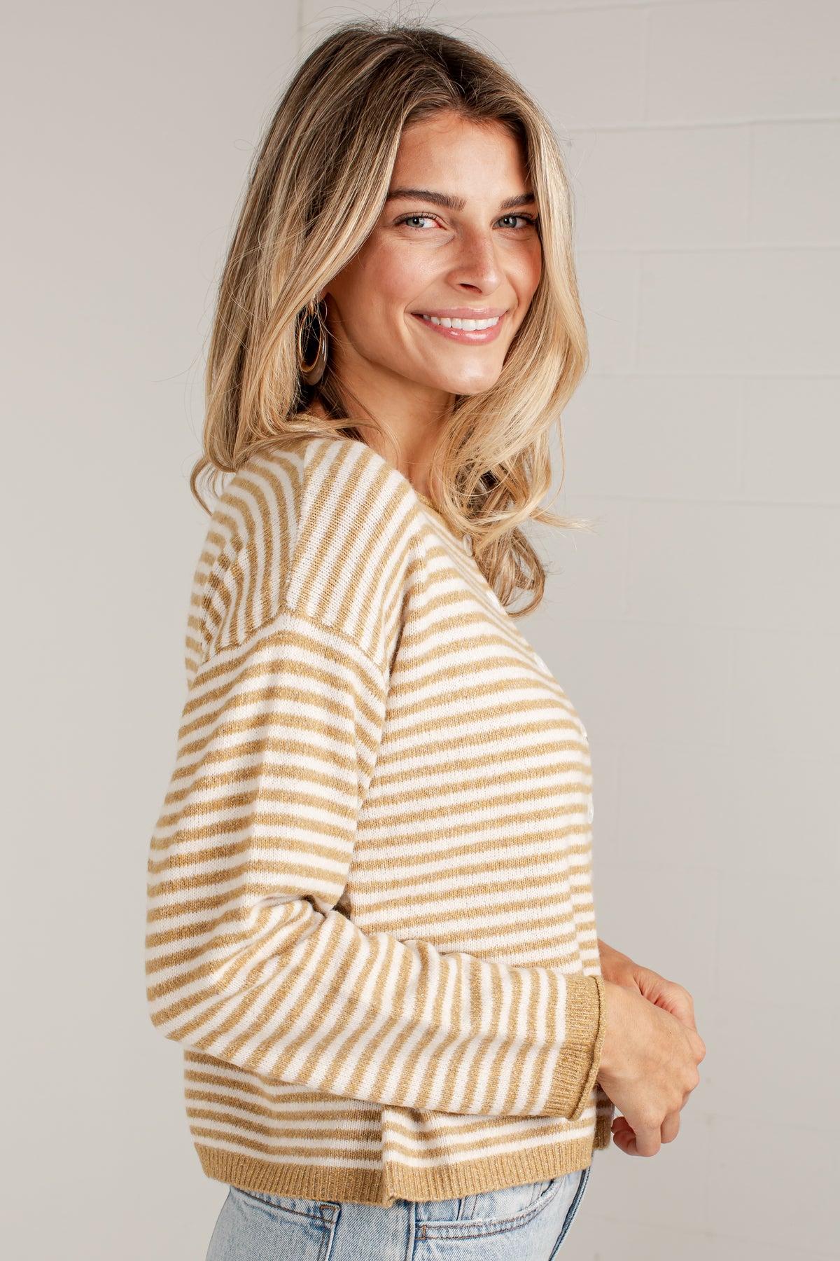 A woman wearing a beige and white horizontally striped knit sweater, paired with light blue jeans, photographed against a neutral light gray background.