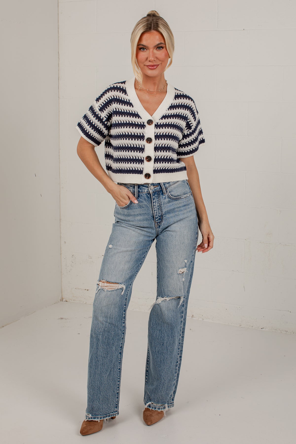 A woman modeling a short-sleeve knit cardigan with navy and white horizontal stripes, paired with light-wash distressed jeans and brown pointed-toe heels, standing against a white studio wall.