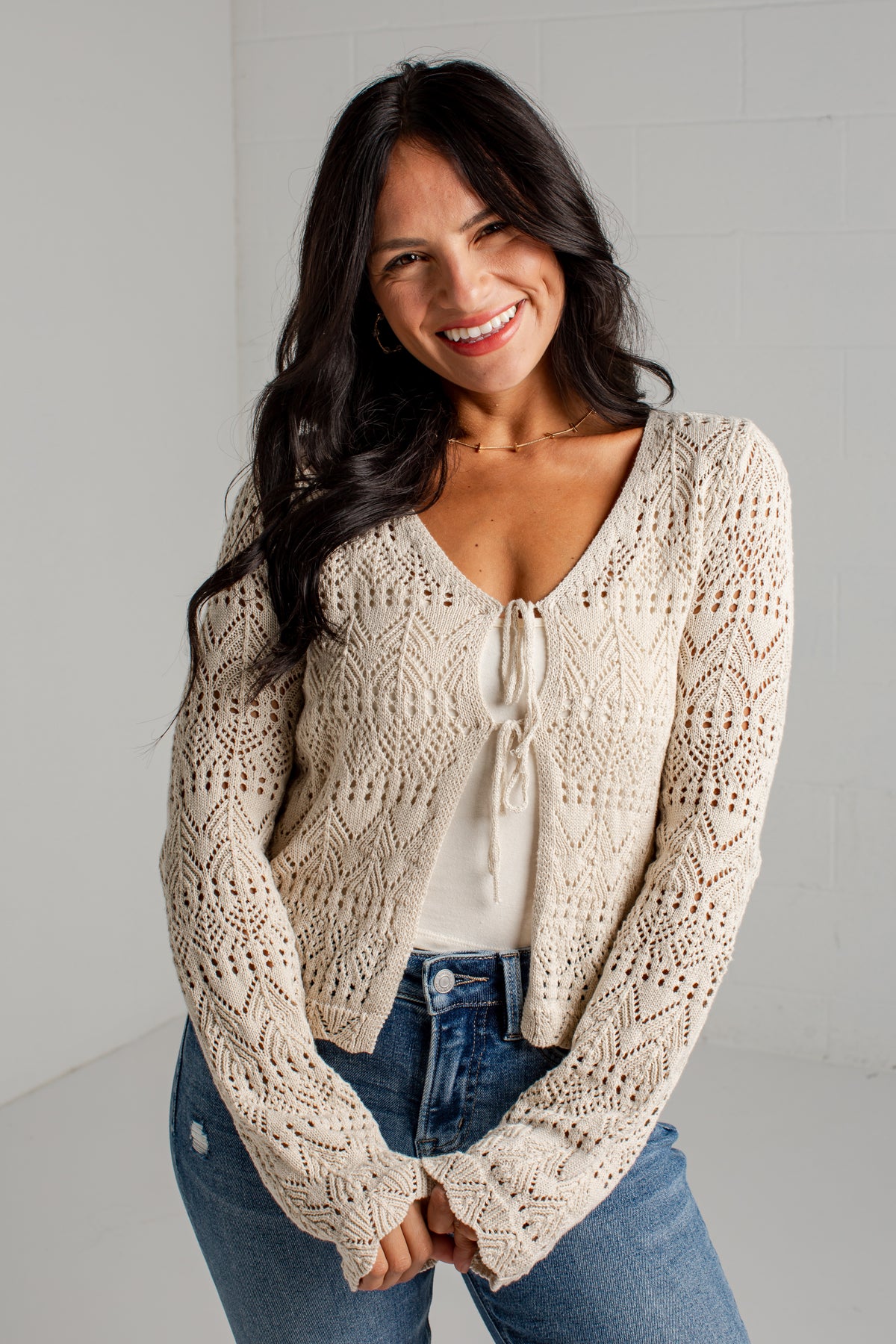 A woman wearing a cream-colored, long-sleeve open-knit cardigan with a tie-front detail, styled over a white top and blue jeans.