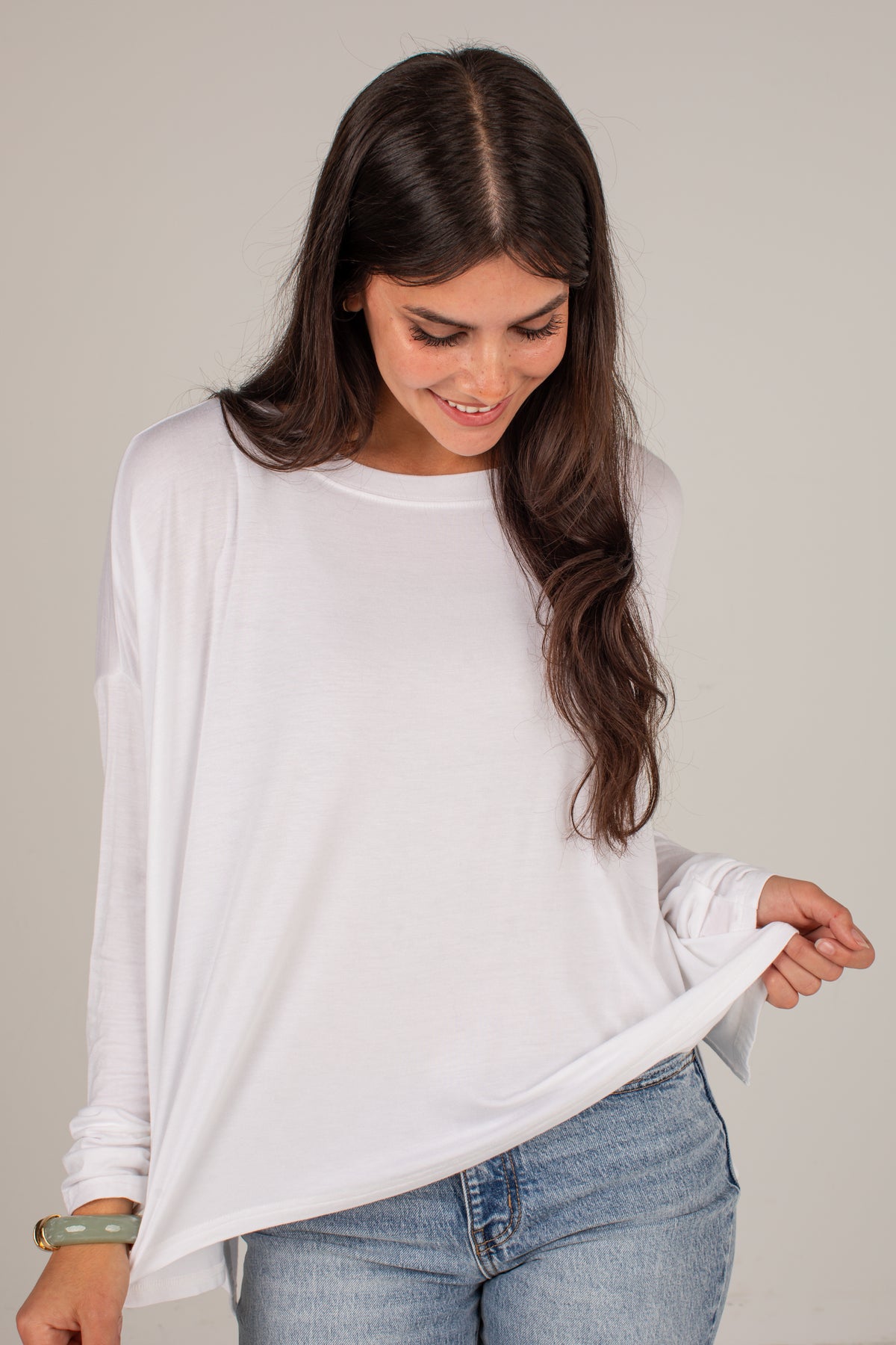 A woman wearing a white long-sleeve crewneck top and light blue jeans, holding up the hem of the shirt against a plain light gray background.