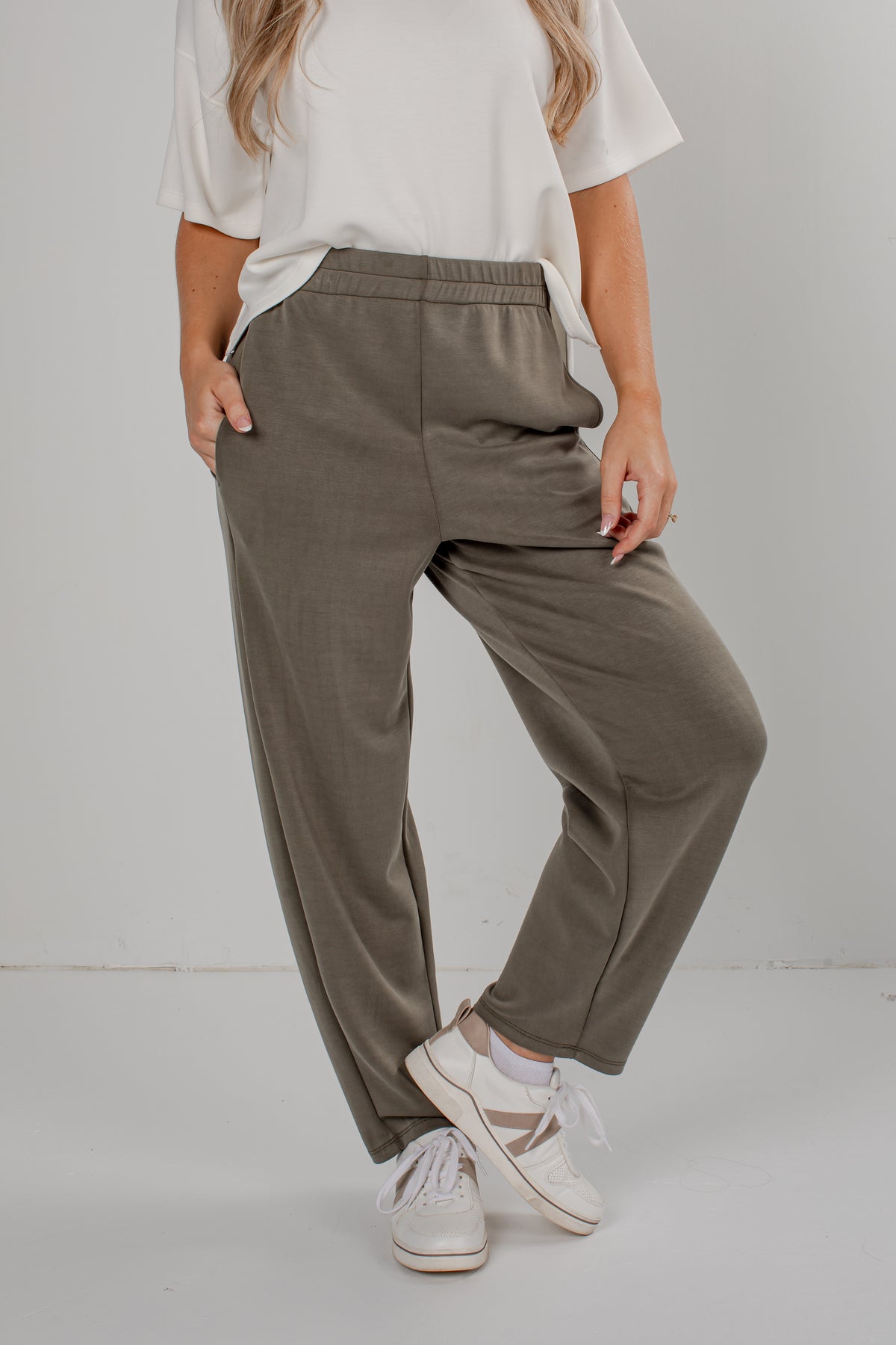 A person wearing loose, wide-leg olive green pants with an elastic waistband, styled with a white t-shirt and white sneakers against a plain light gray studio background.