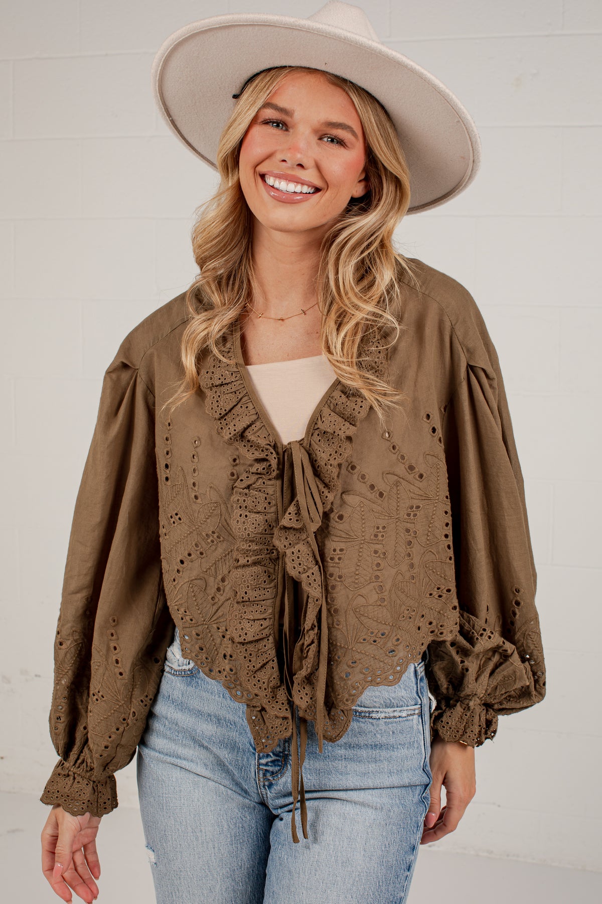A woman wearing a brown eyelet lace blouse with ruffles and long puff sleeves, paired with a wide-brim beige hat and light blue jeans, standing against a white brick wall.